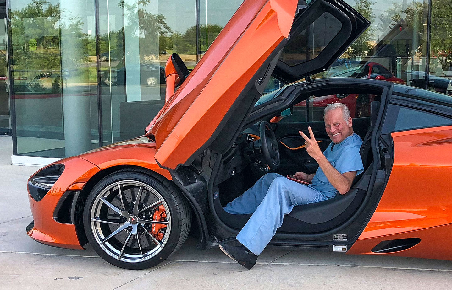 Dr. Billy (Dr. WIlliam Cohn) in blue scrubs sitting in an orange McLaren sports car with its scissor door open, smiling and flashing a peace sign in front of a glass-walled dealership.
