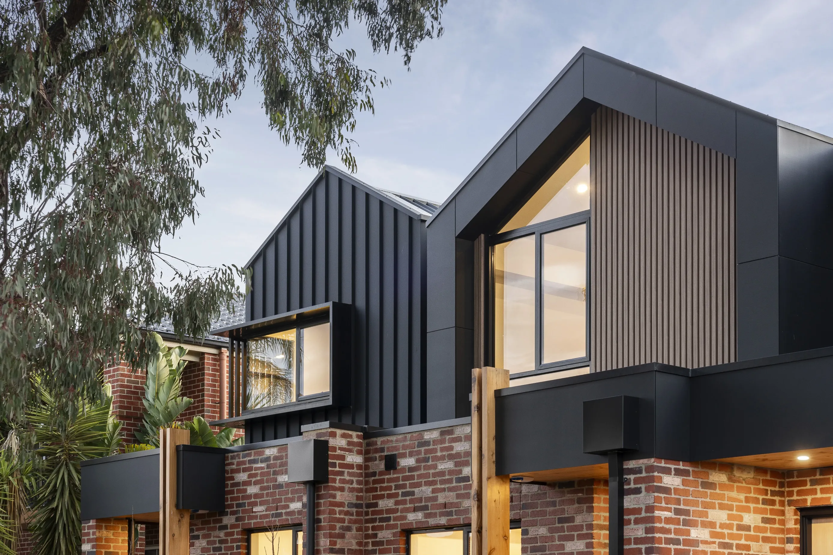 Upper-level detail of the Coburg townhouses at dusk with black metal-clad gables, timber-batten panels, recycled-brick walls, timber posts and warmly lit windows among gum trees