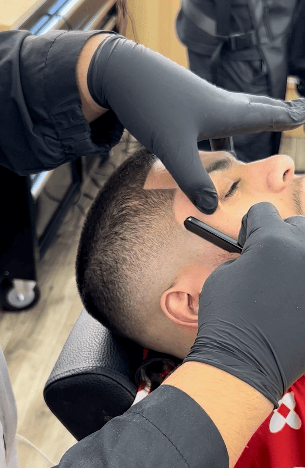 A barber in black gloves uses a straight razor to shape a client’s hairline during a haircut.