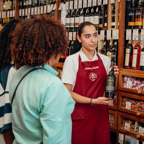 一名身穿红色围裙的店员手拿一瓶葡萄酒，与两位顾客交谈。背景是摆满葡萄酒的货架。