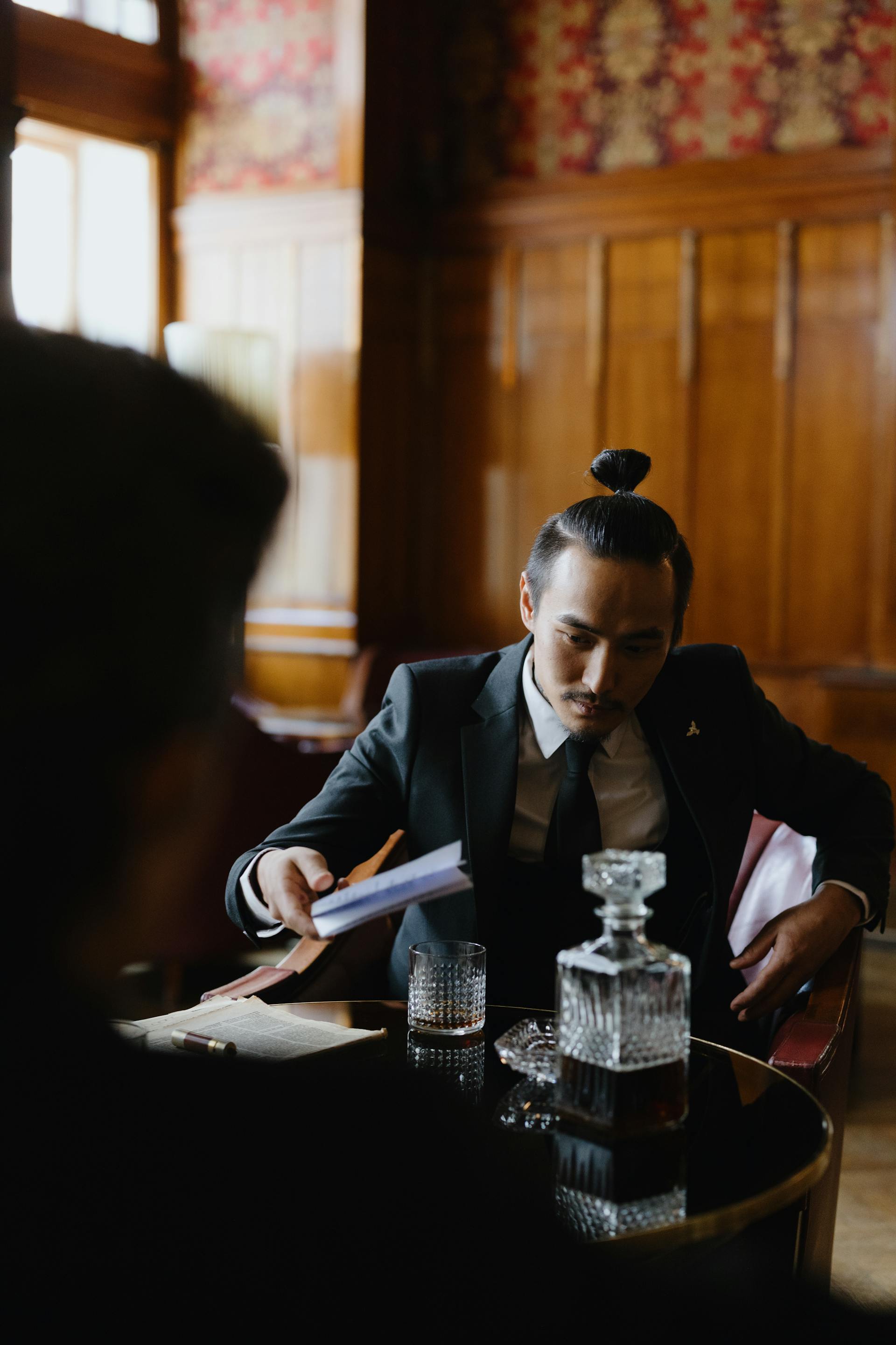 A man with his hair in a top knot, wearing a black suit and tie, leaning forward in a leather chair to slide a small envelope across a table toward another person.