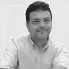 Smiling person sitting at a desk, wearing a sweater, with a drink and food in front of him.