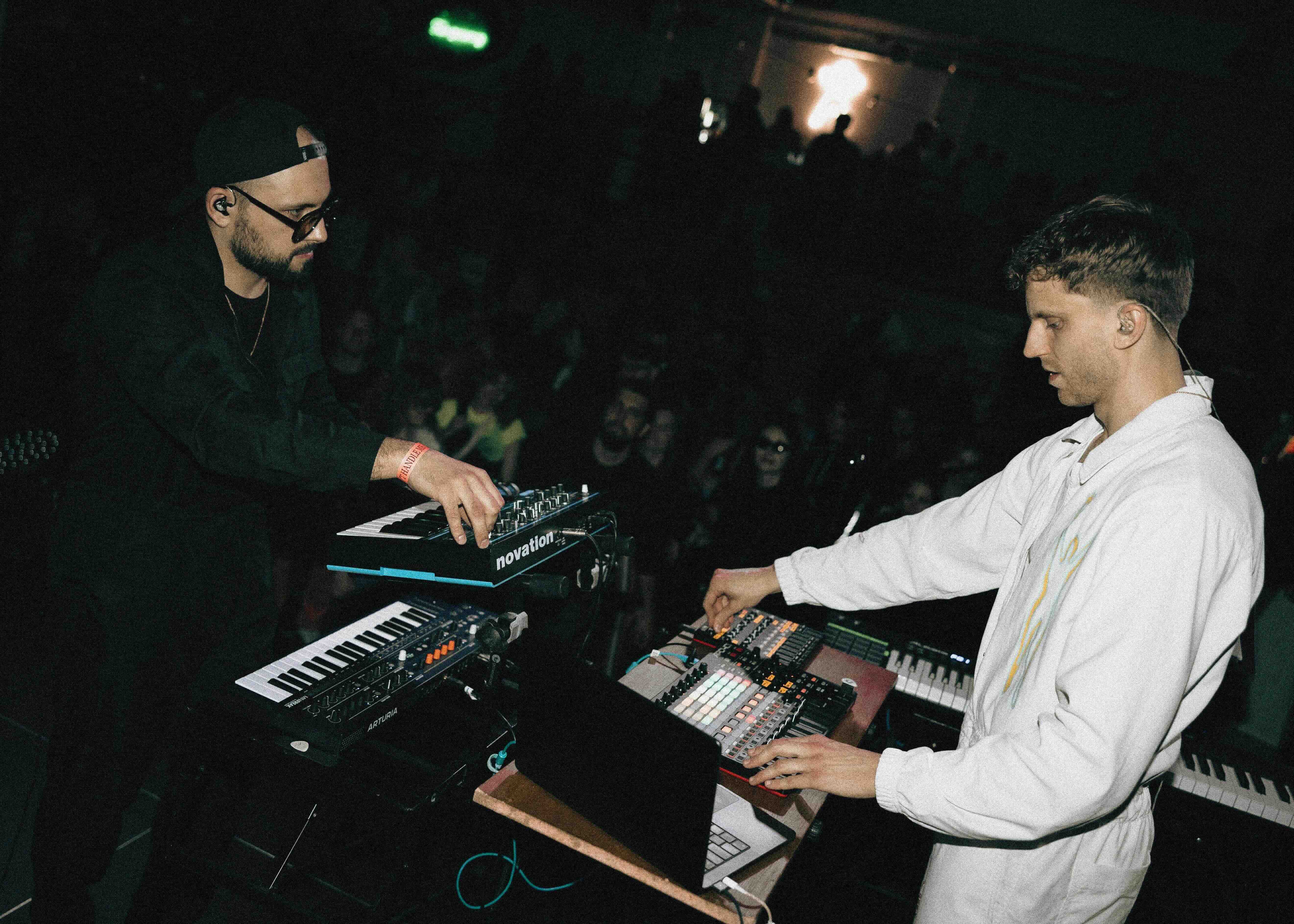 Two musicians performing behind synthesizers on a teal-lit stage