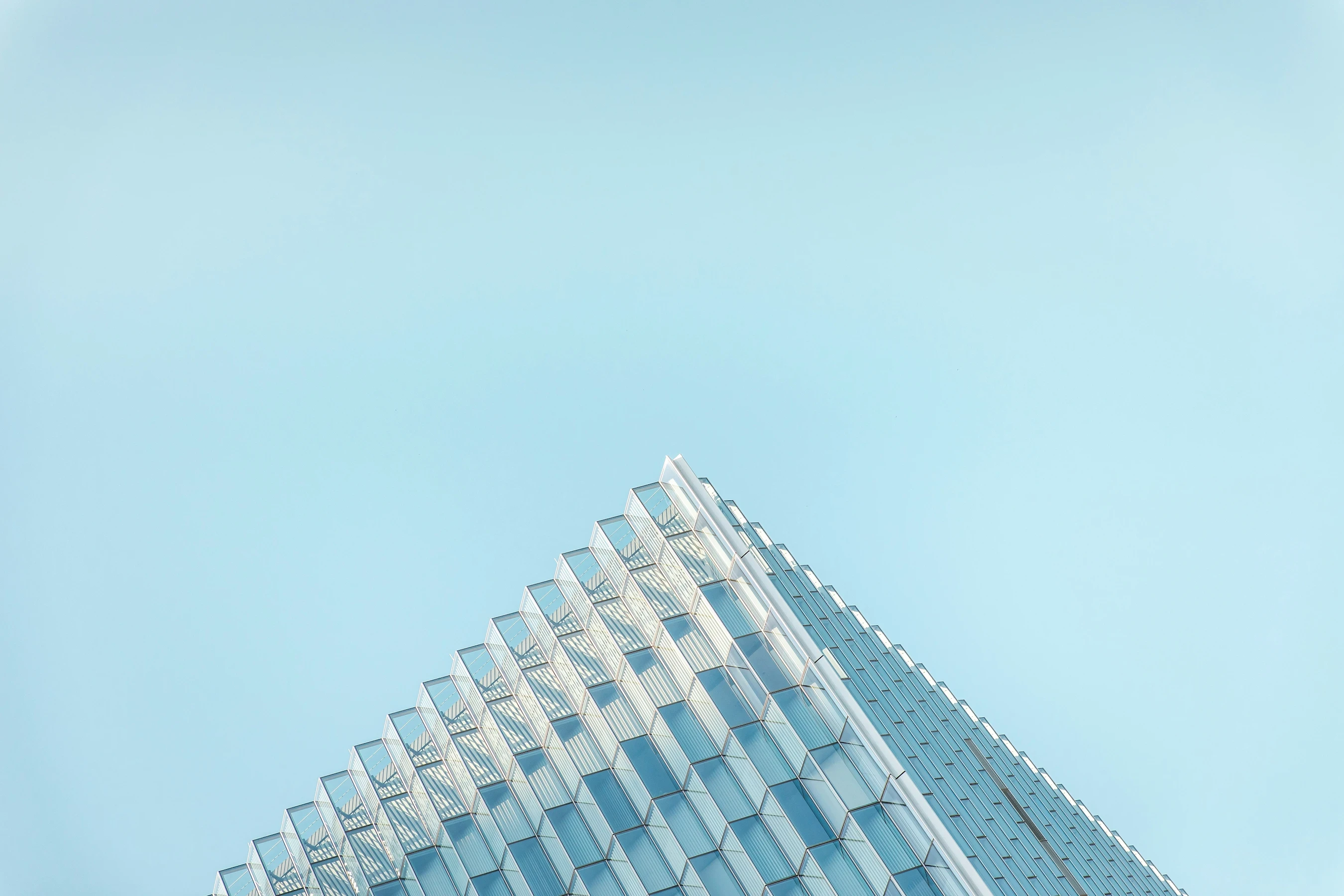 Low-angle shot looking up at the sharp, triangular corner of a modern glass skyscraper against a clear, pale blue sky. The geometric windows form a clean grid pattern.