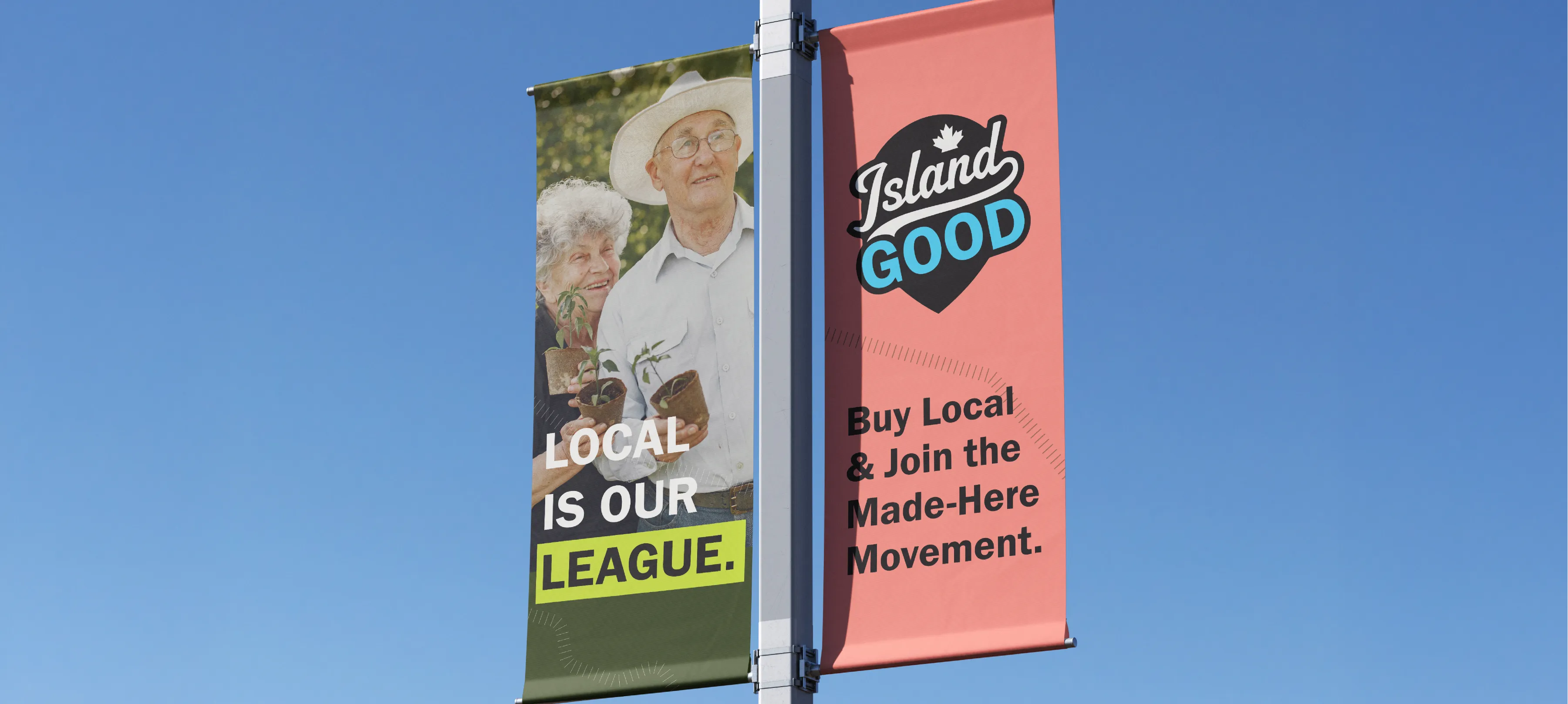 two street banners hangining on a lamp post in front of blue sky. The first banner has "Local is our league" printed on it with "league" highlighted with a lime green box with an older couple in the background holding plants. The second banner is coral and has the Island Good logo with the text "Buy Local & Join the Made Here Movement" written below.