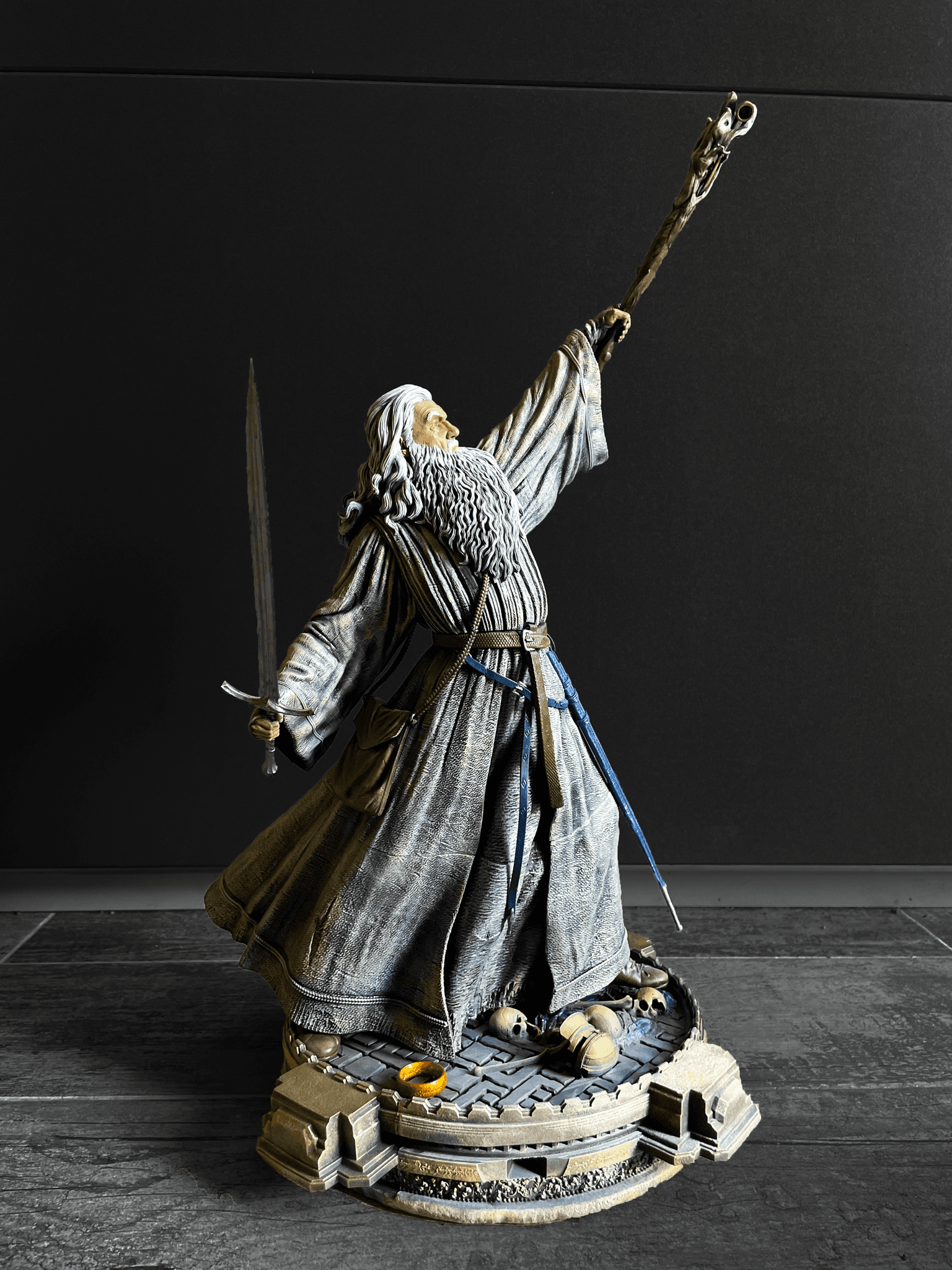 Ručně malovaná figurka Gandalfa z Pána prstenů, držící hůl a meč, na podstavci s lebkami a Prstenem