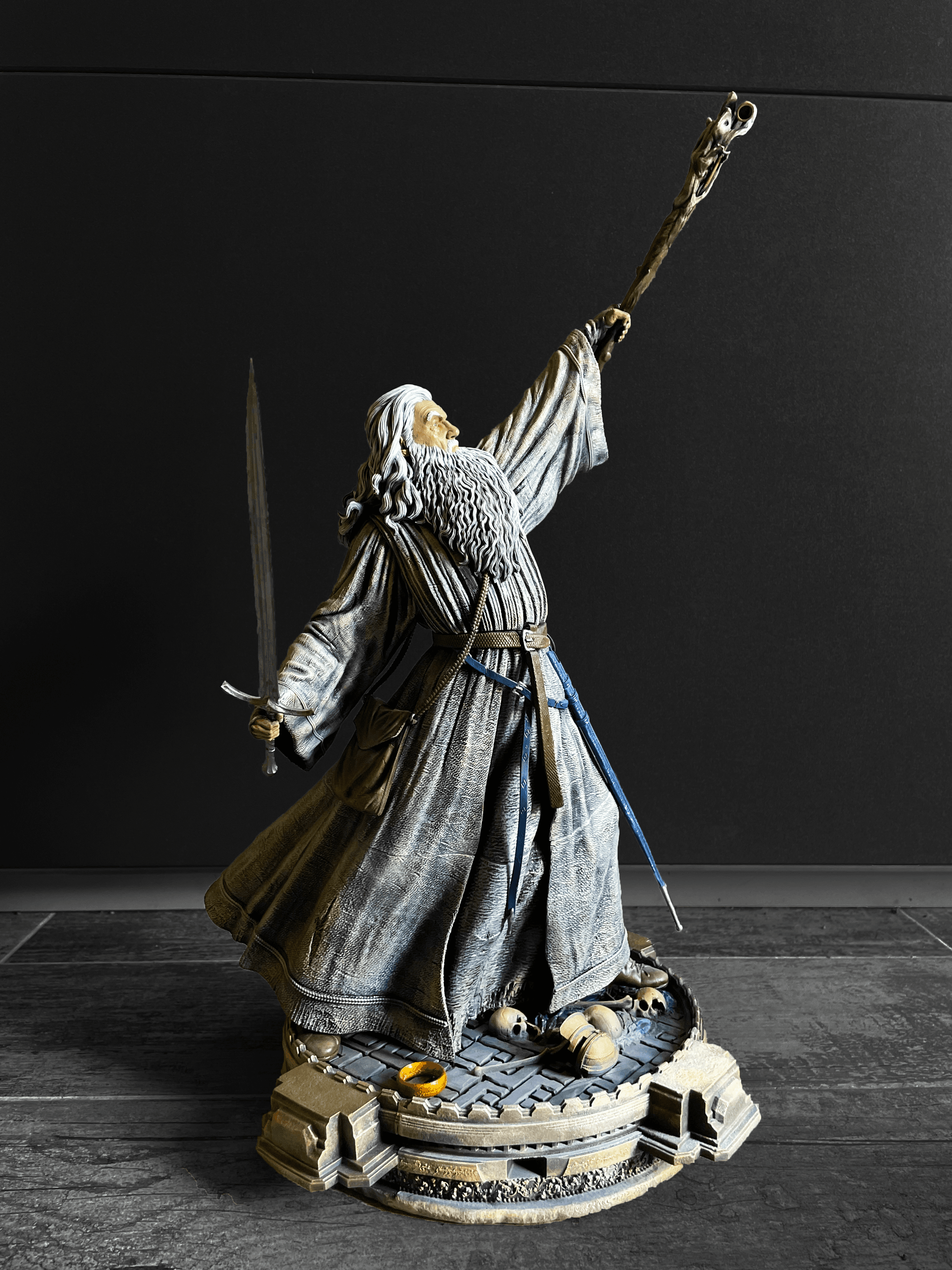 Ručně malovaná figurka Gandalfa z Pána prstenů, držící hůl a meč, na podstavci s lebkami a Prstenem