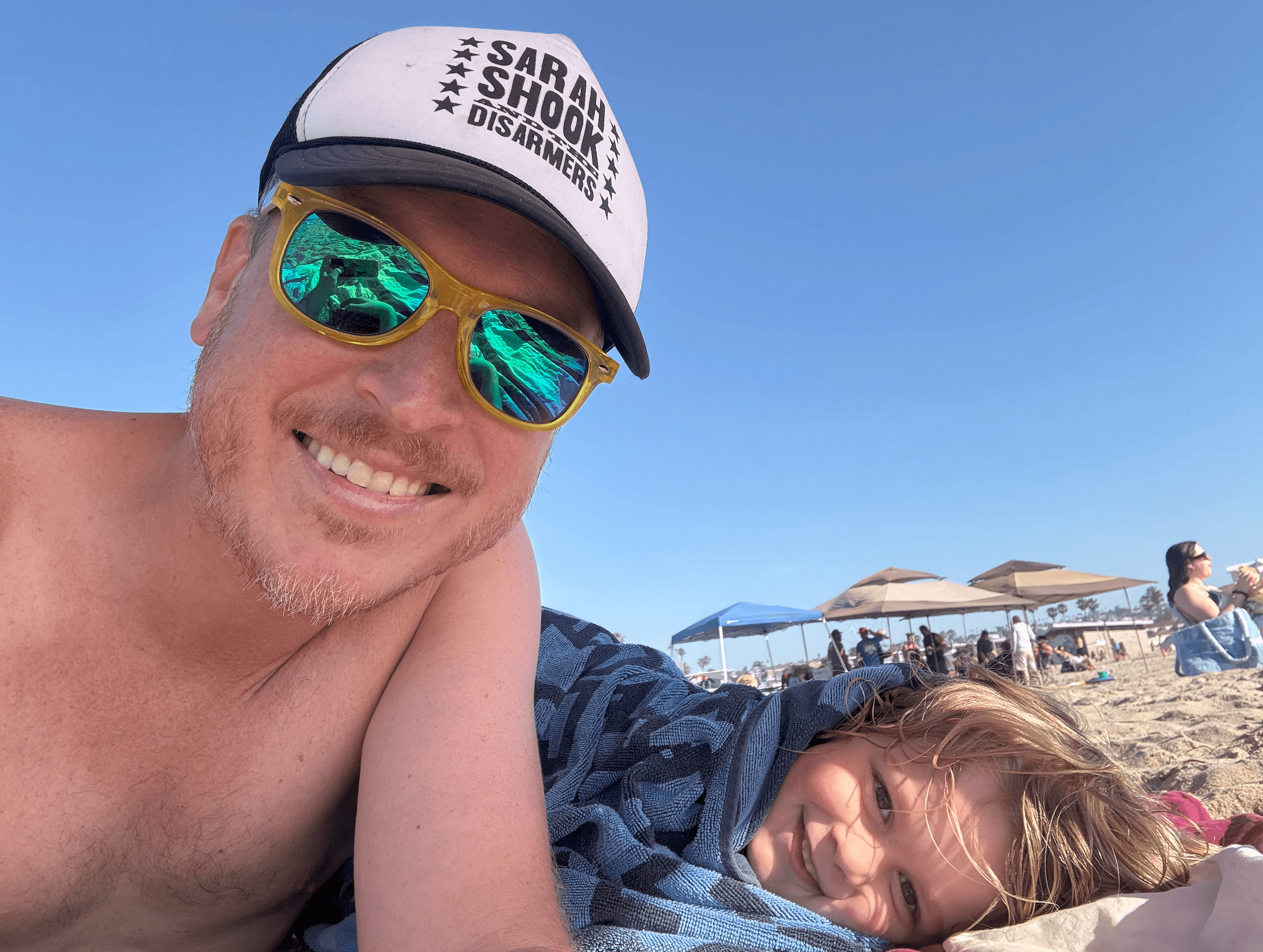 A young person with curly hair smiles at the camera while sitting in front of a laptop against a dark background.