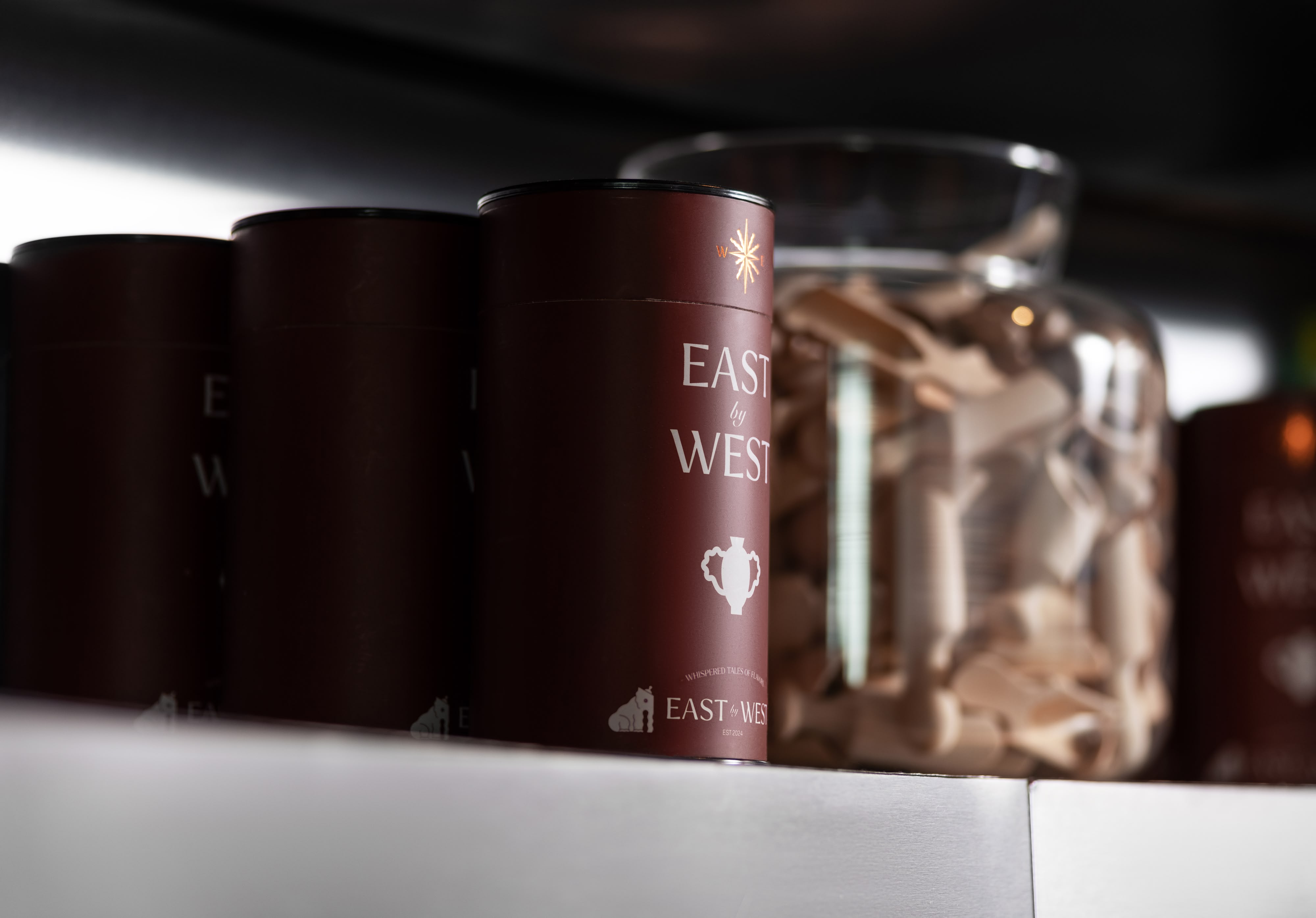 East by West restaurant maroon cylindrical containers with star and teapot icons on labels, displayed on shelf beside glass jar of wooden sticks.