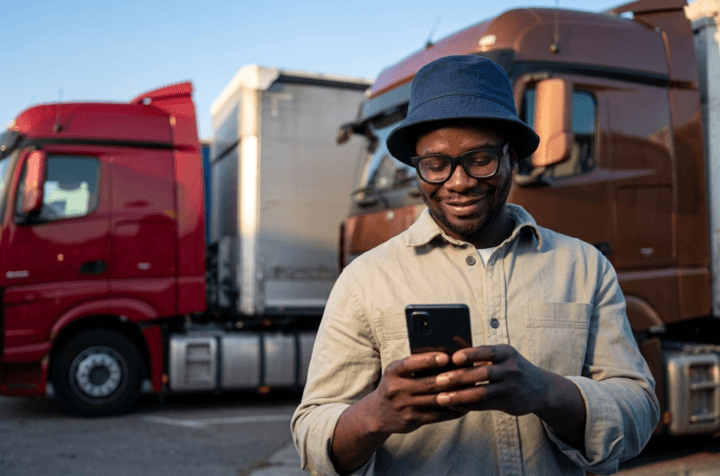 A truck driver in a bucket hat and glasses stands smiling at his phone in a truck yard, with a red and a brown semi-truck parked behind him, using the free load board app to search for loads.