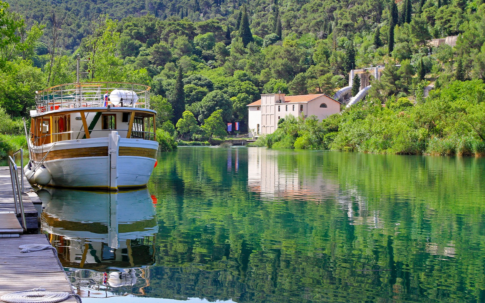 Barco atracado en el río del Parque Nacional Krka, con exuberante vegetación y un edificio histórico de fondo.