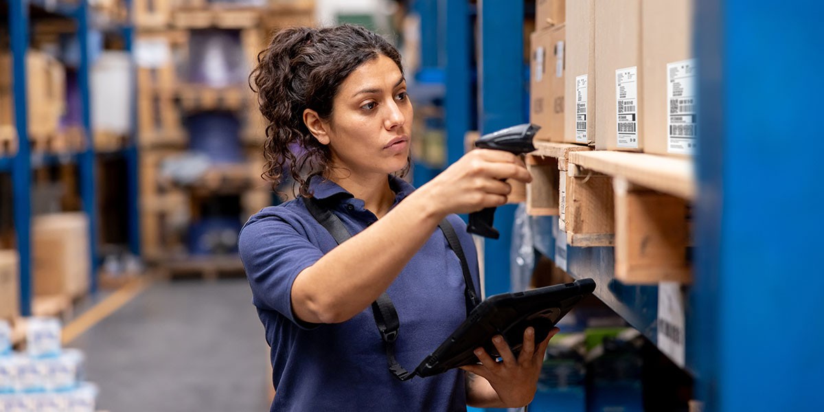 A woman working in a warehouse, checking inventory on a clipboard while organizing shelves in the background.