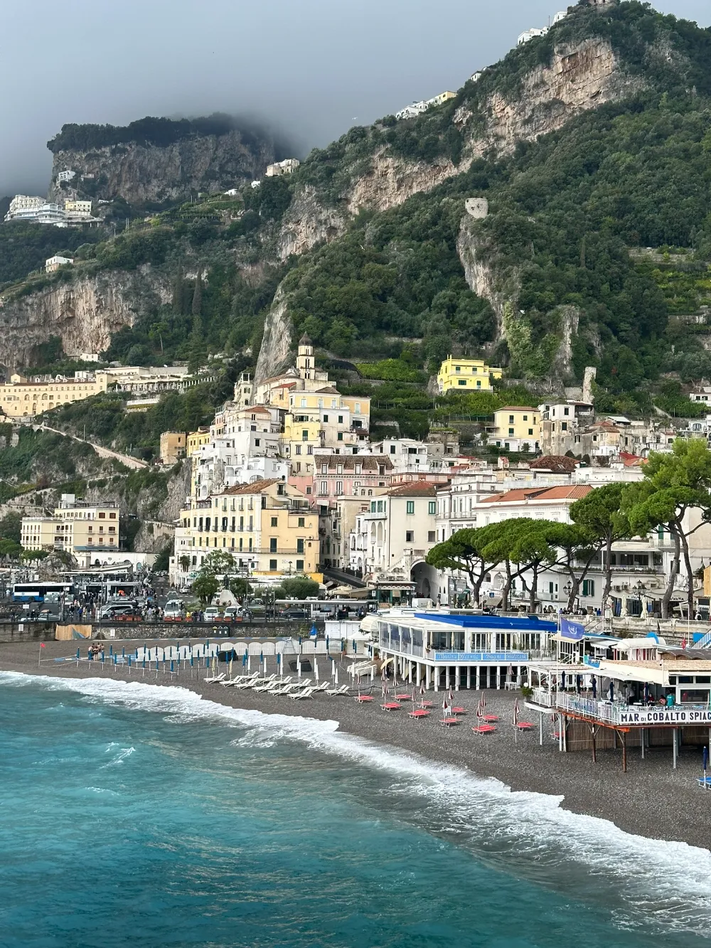 Amalfi town and its beach seen from the cliffs above, with turquoise water and pastel buildings.