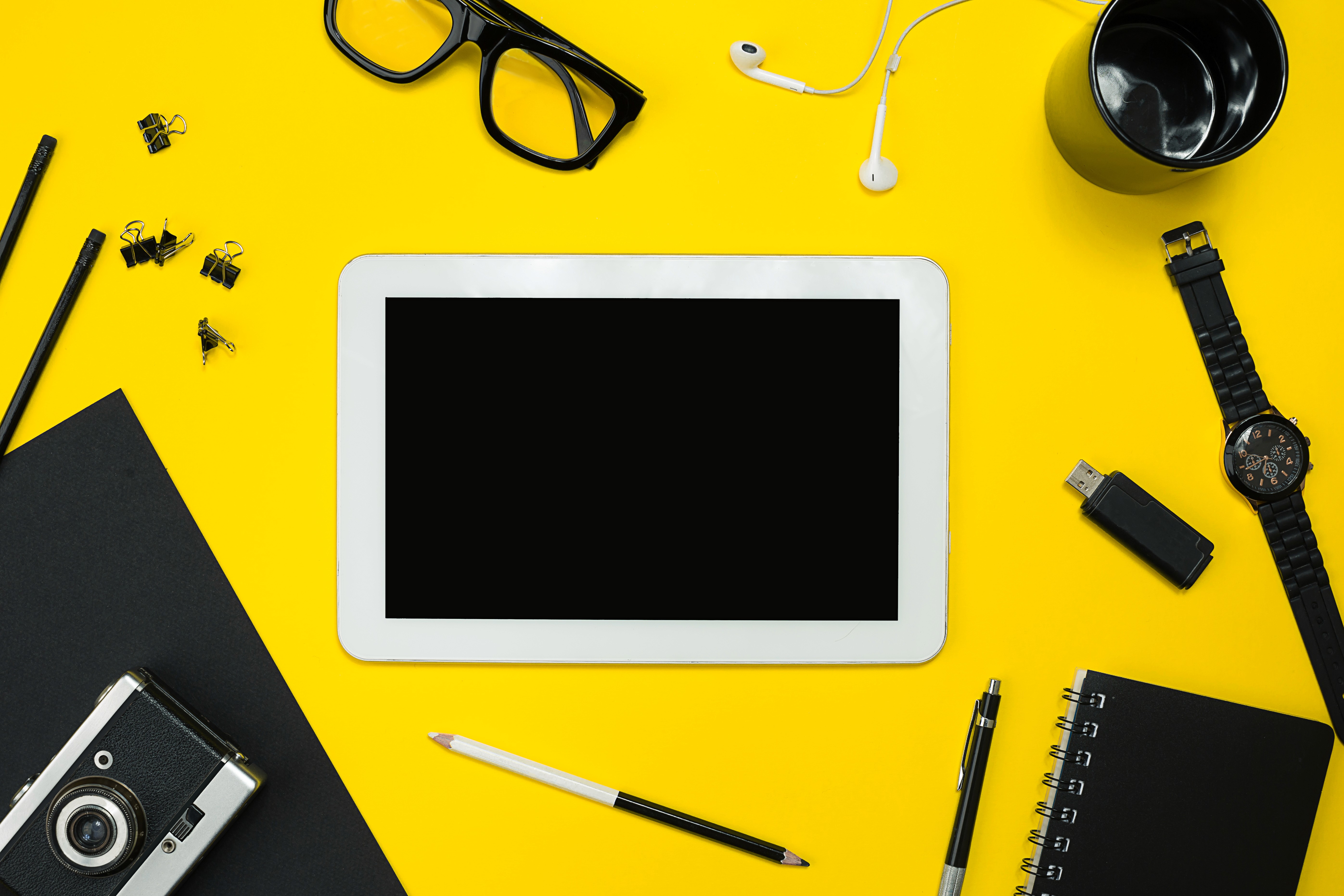 Top-down view of a white digital tablet with a blank black screen centered on a bright yellow background, surrounded by workspace essentials like eyeglasses, earbuds, a pen, and a notebook for a modern tech mockup.