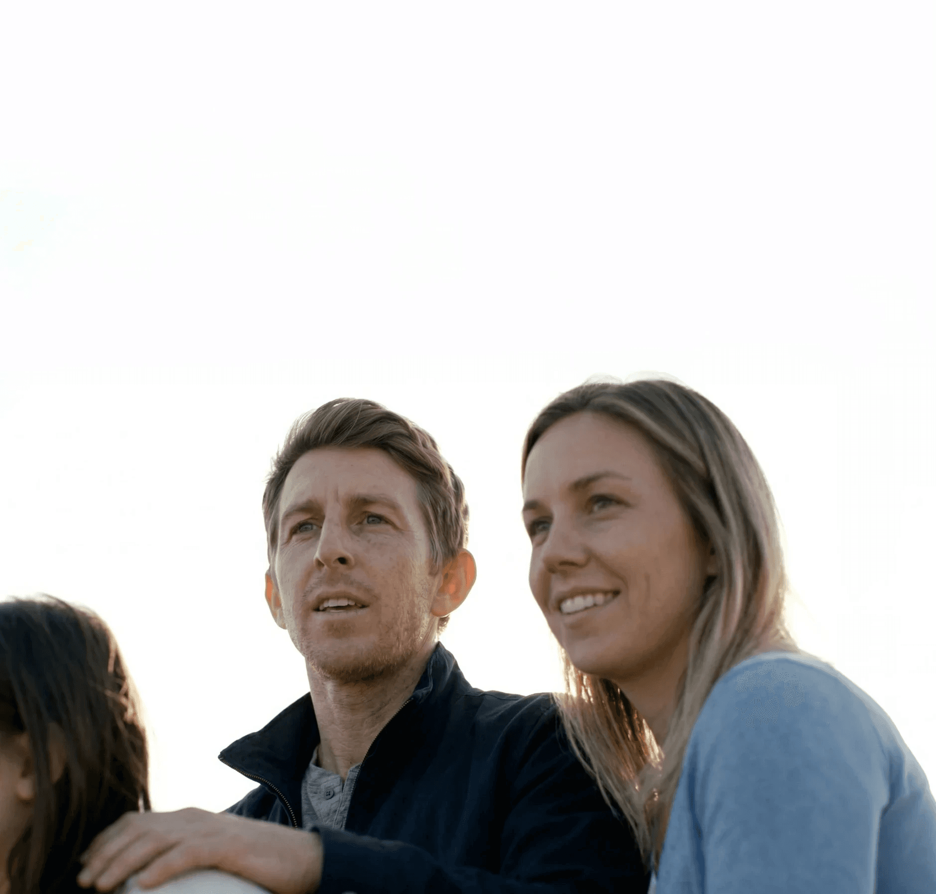 A family stands closely together outdoors under a bright sky, looking content and focused on something in the distance.