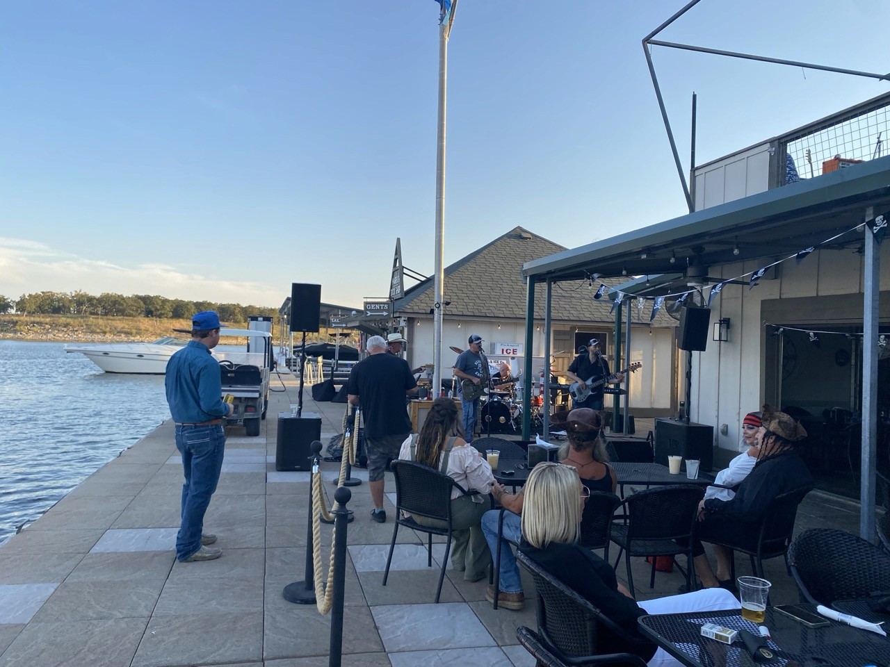 A small group of people enjoy live music by a band performing outdoors on a dockside patio under a clear sky, with a scenic waterfront view in the background.
