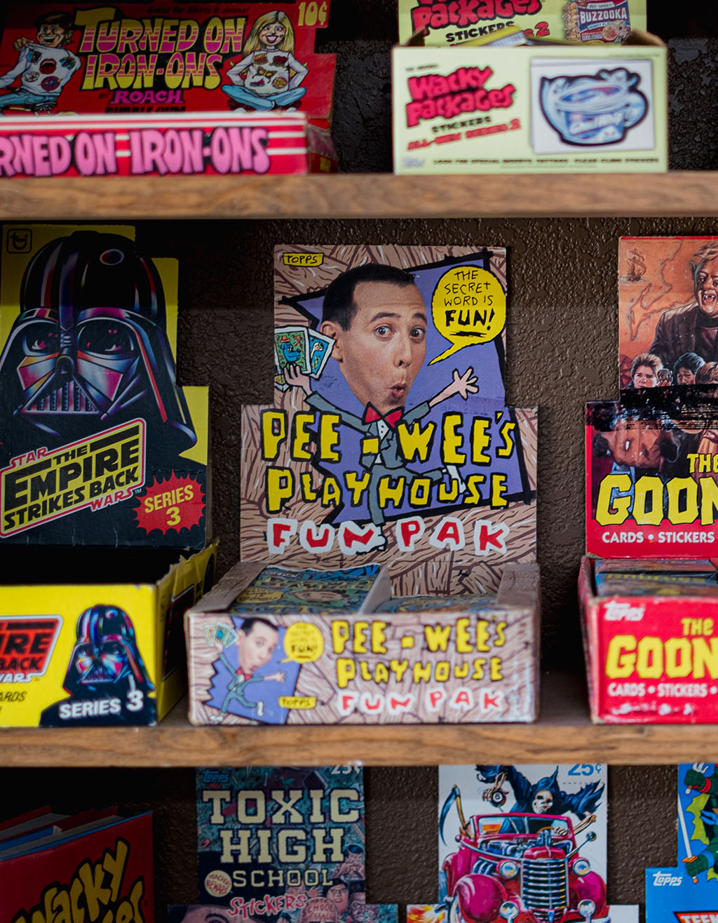 Close-up of vintage sticker pack boxes on a shelf, featuring Pee-wee’s Playhouse Fun Pak centered among classic packs like Star Wars, The Goonies, and Toxic High School.