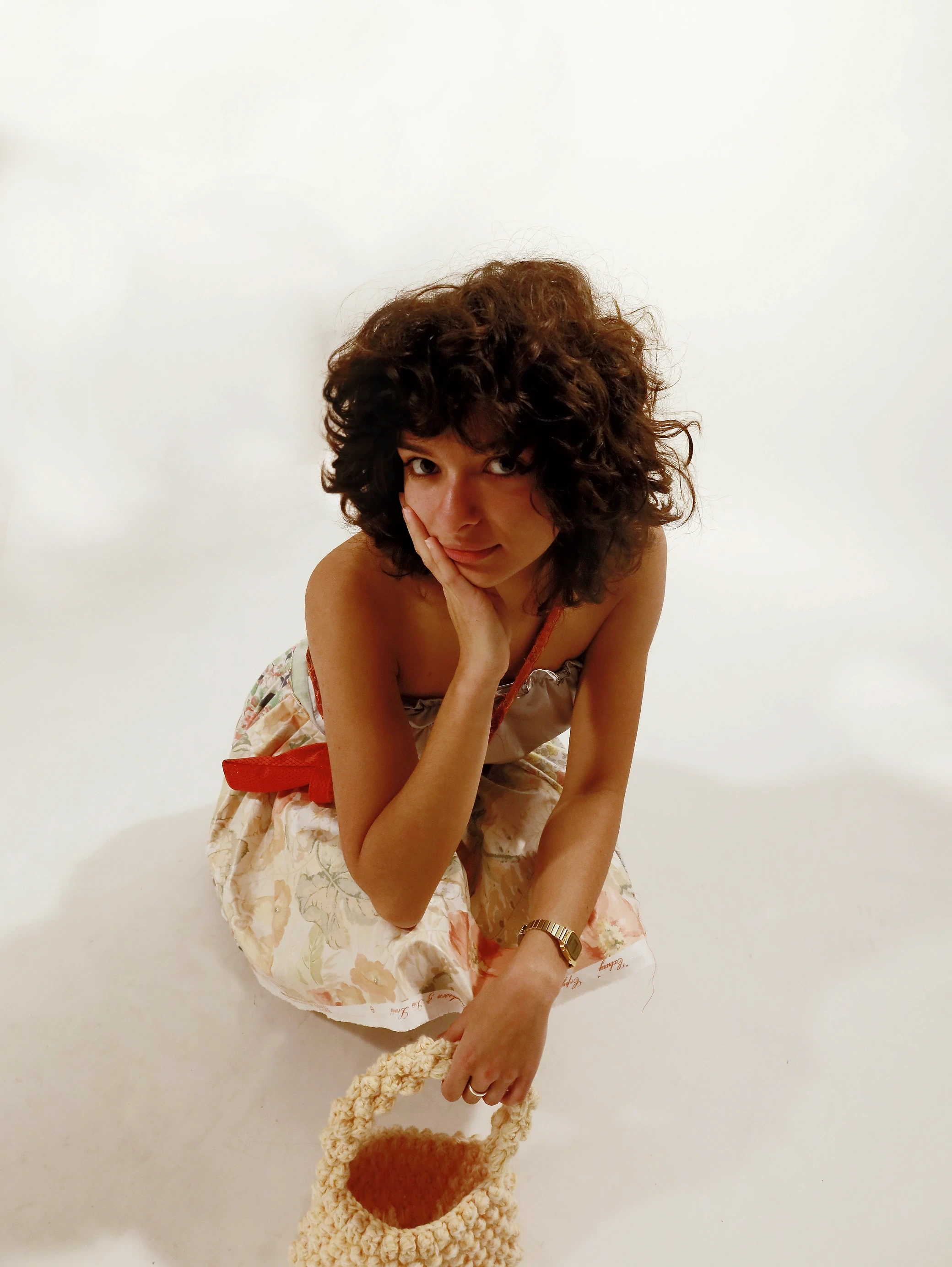 A person with curly hair sits, looking thoughtful, wearing stylish casual attire against a light background.