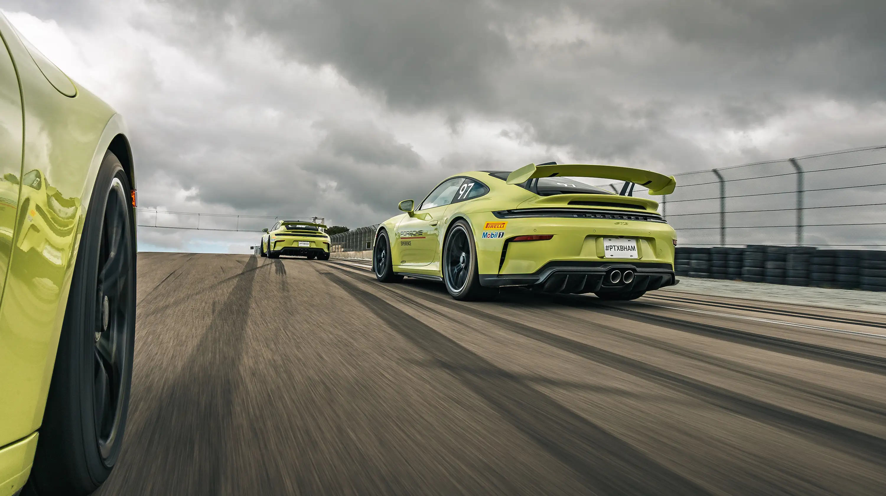 Low-angle view of yellow Porsche track cars driving on a racetrack under dark, stormy clouds.
