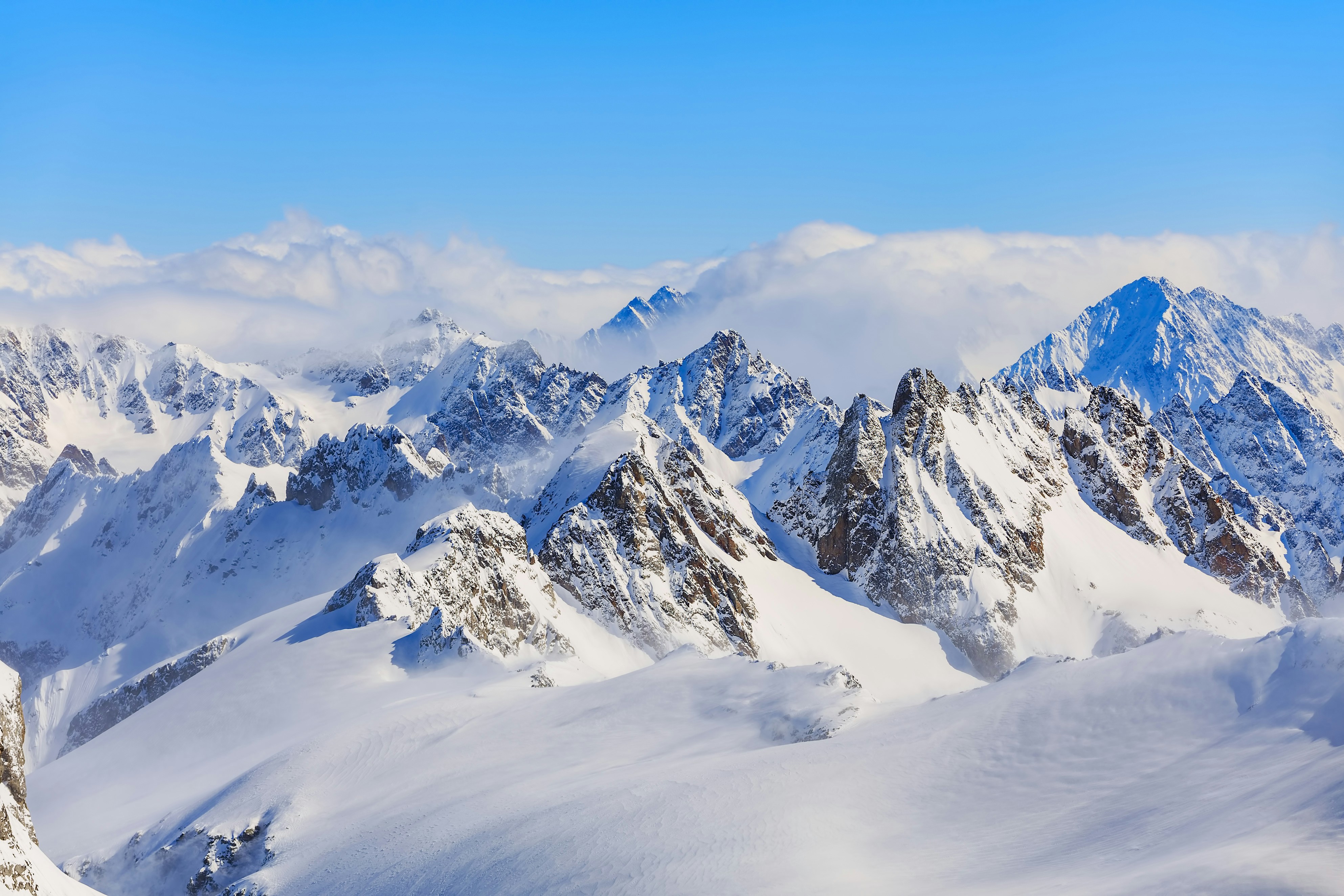 Snow-covered mountain peaks with scattered clouds against a clear blue sky.