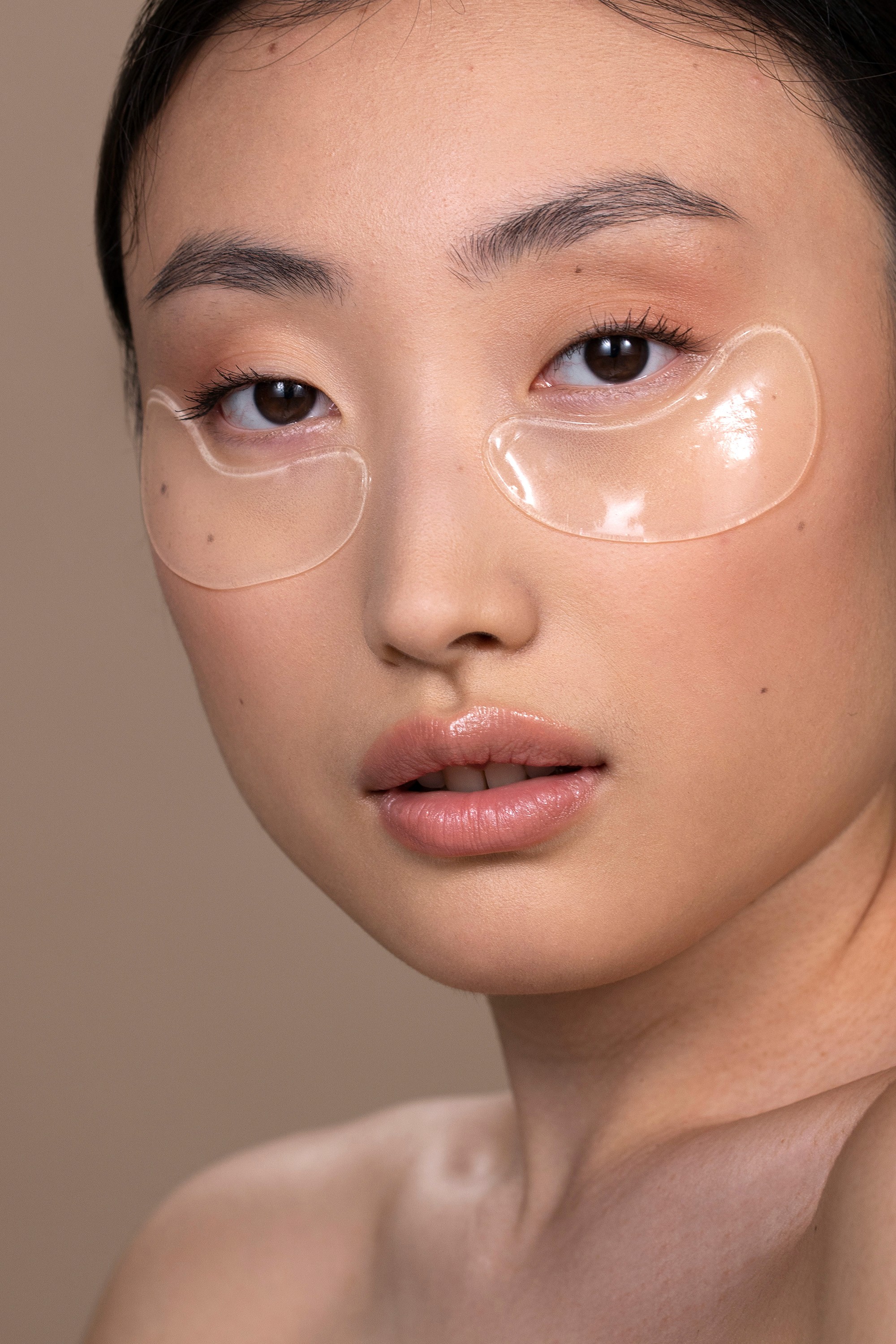 Close-up of a woman with under-eye gel patches, natural makeup, and smooth skin, against a neutral beige background