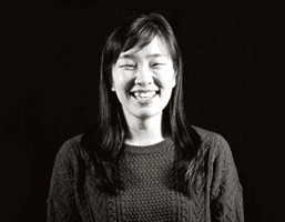 A black and white portrait of a woman with long dark hair. She is smiling and wearing a dark knitted sweater with a textured pattern. The background is solid black.