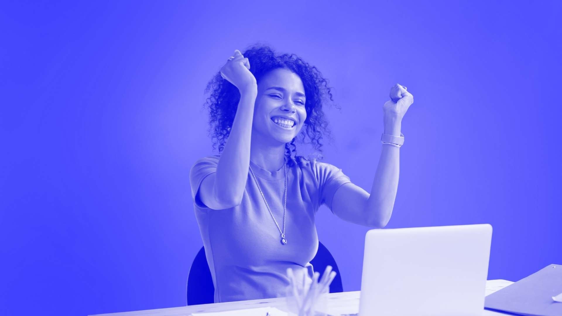 A woman smiling and fist bumping the air while sat in front of her deak and laptop