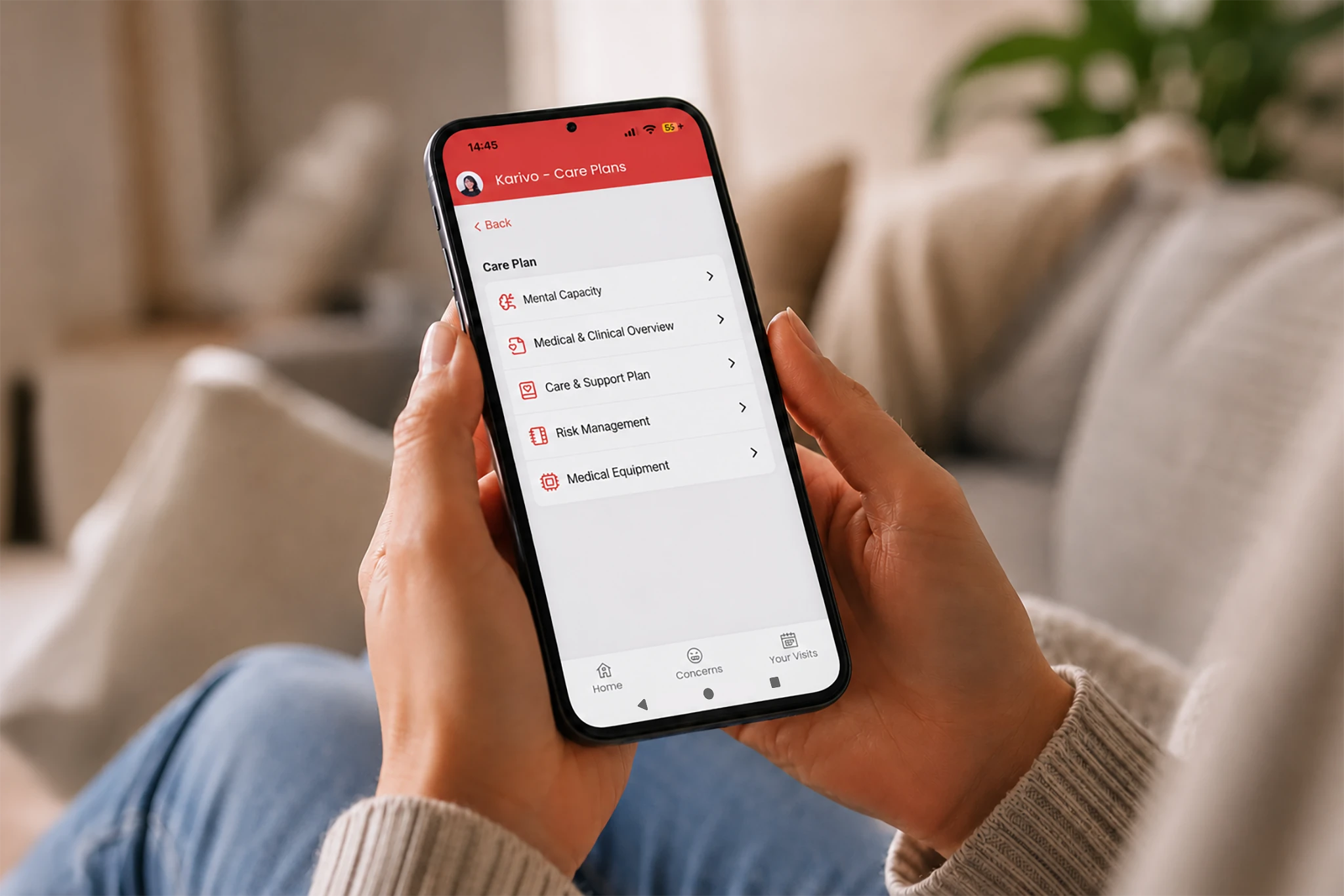 Close-up of a person holding a smartphone at home, displaying the Karivo Care Plans app with options for mental capacity, clinical overview, care and support, risk management, and medical equipment.