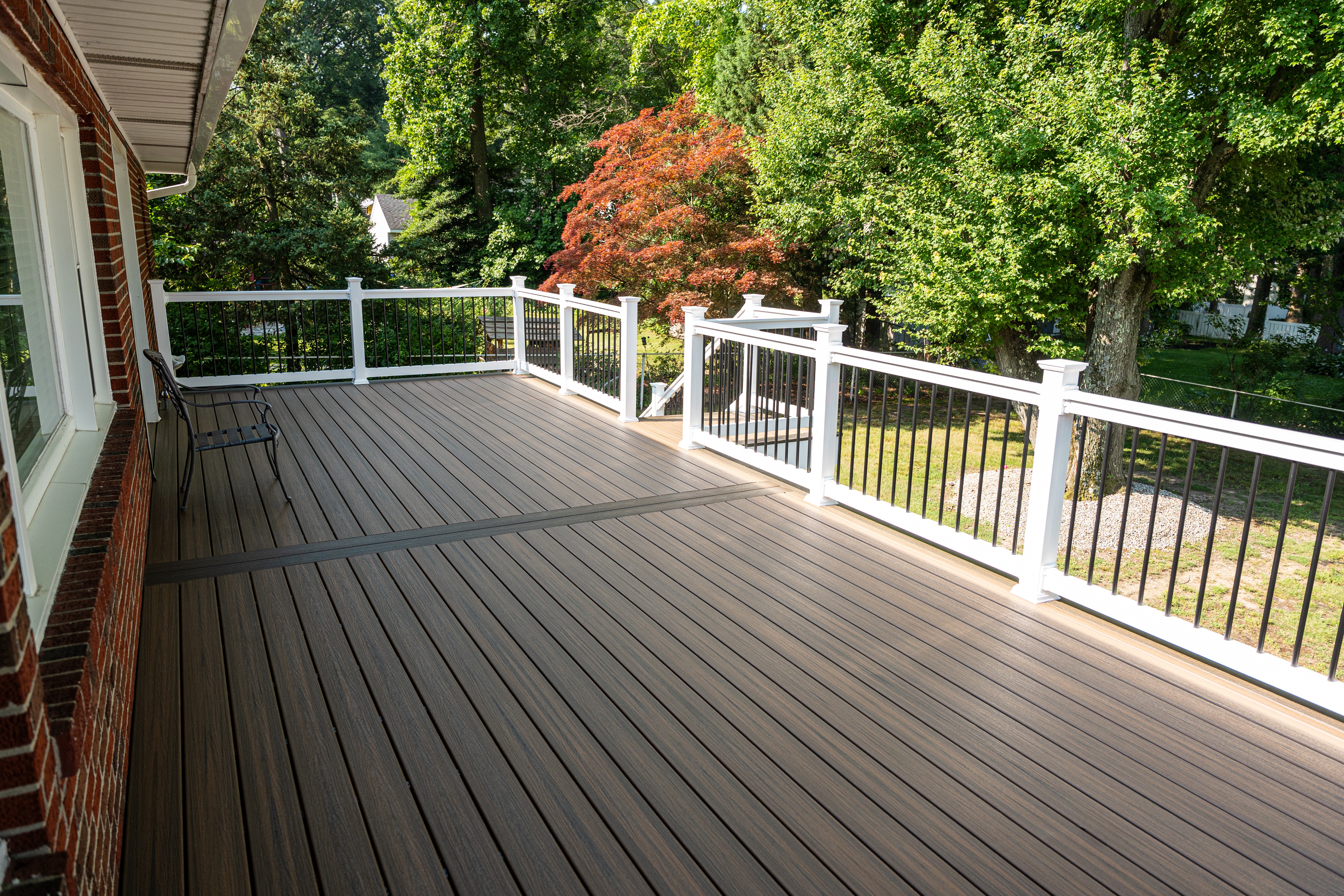 A spacious backyard deck with a wood-look composite surface and white railing overlooks a lush garden with vibrant green and red-leaved trees, providing a serene outdoor setting.