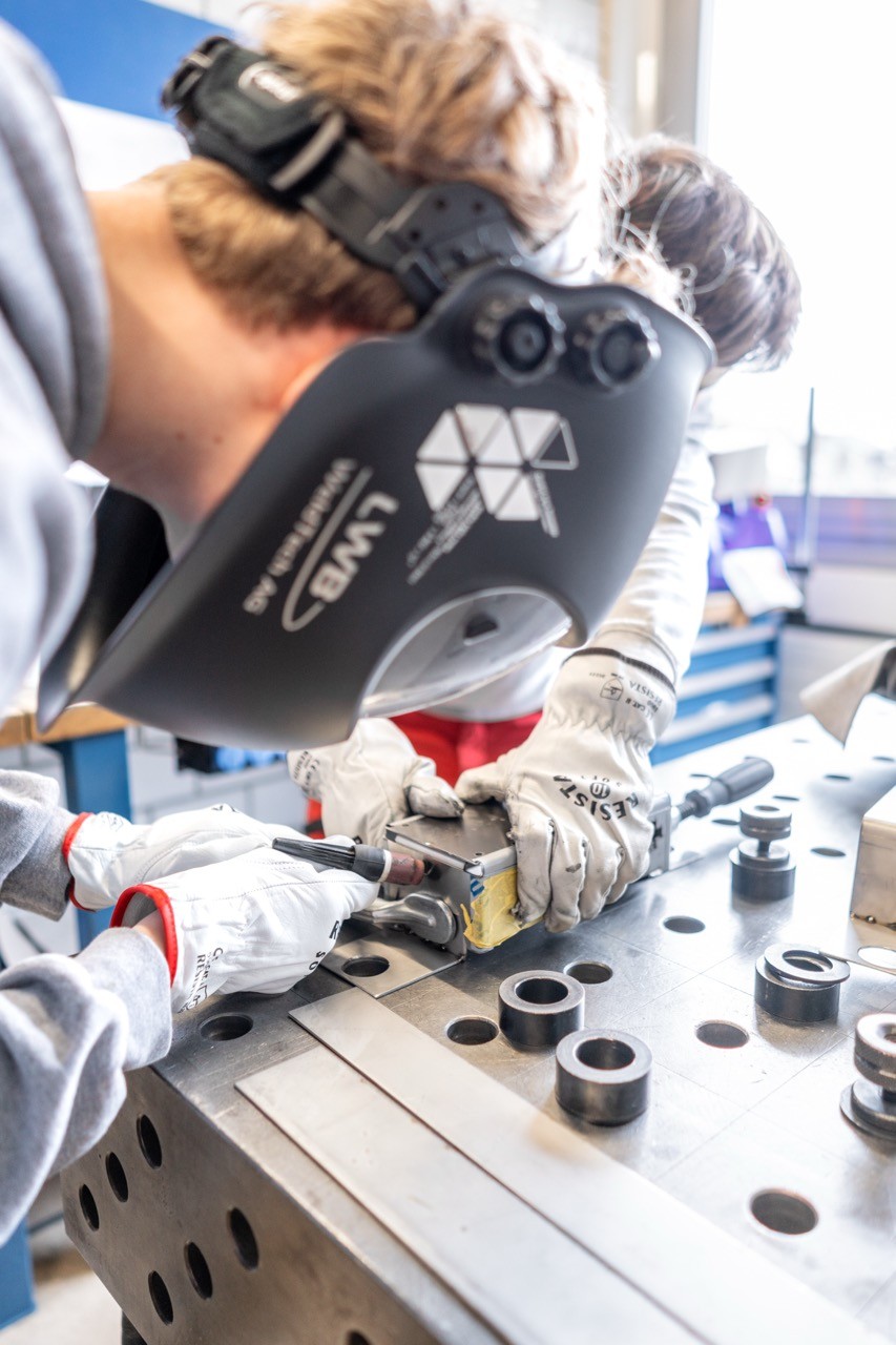 Lernender schleift mit Schutzhelm und Handschuhen ein Edelstahlteil auf der Werkbank