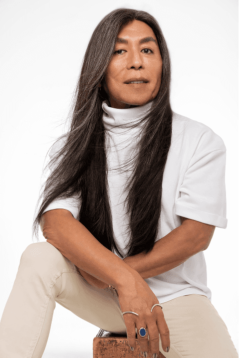 Celso Kamura a sorrir para a câmara, com cabelo longo e liso, a usar camisola preta, em fundo cinzento neutro