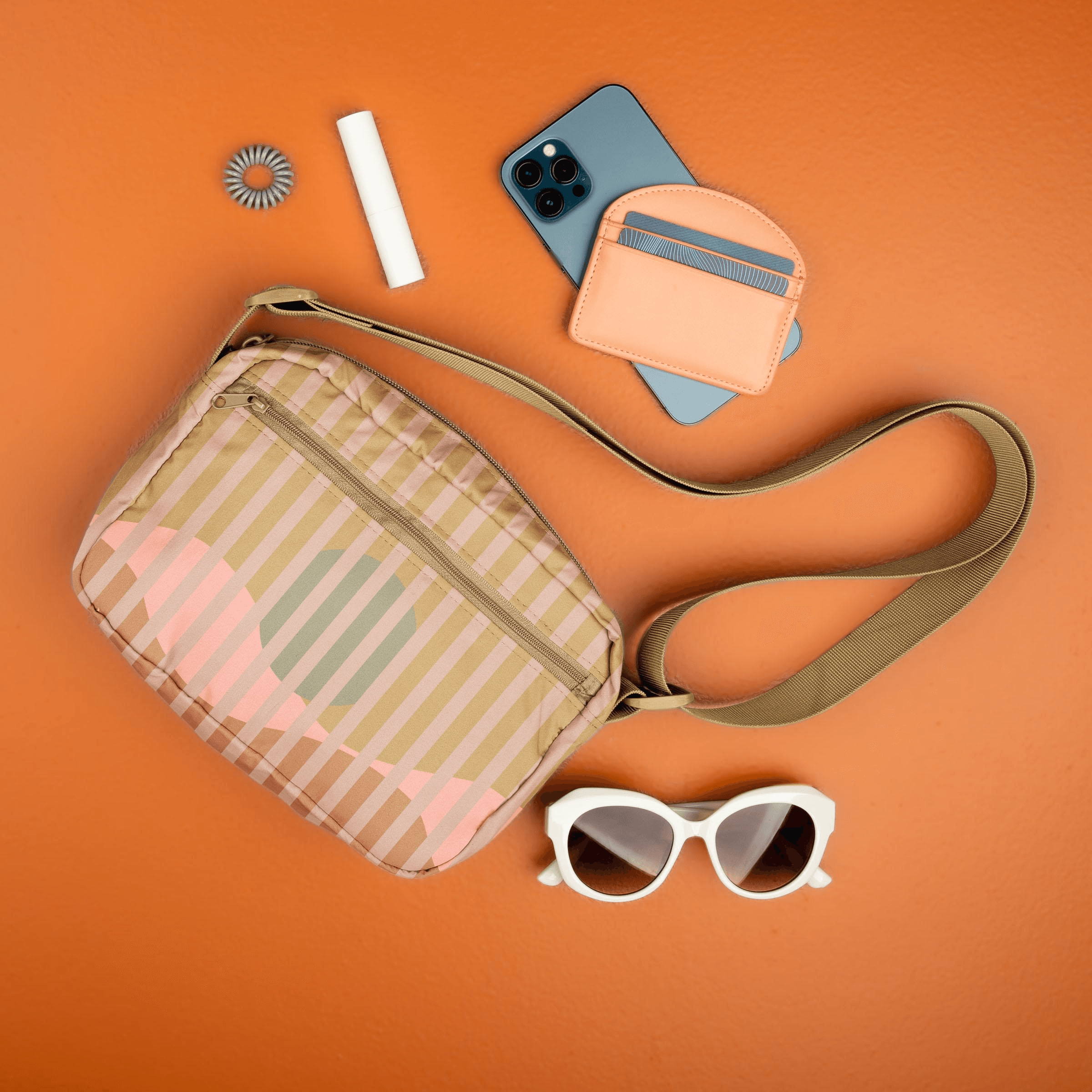 The landscape crossbody tote on an orange back drop with various items such as a phone, wallet, chapstick and sunglasses next to the bag itself.