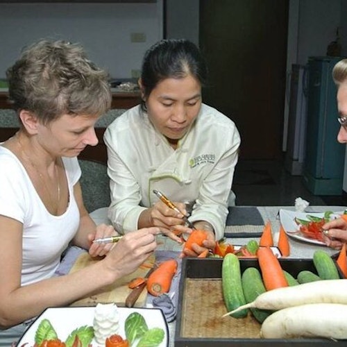 Trei persoane la o masă sculptează legume; mai mulți castraveți, morcovi și ridichi sunt vizibili.