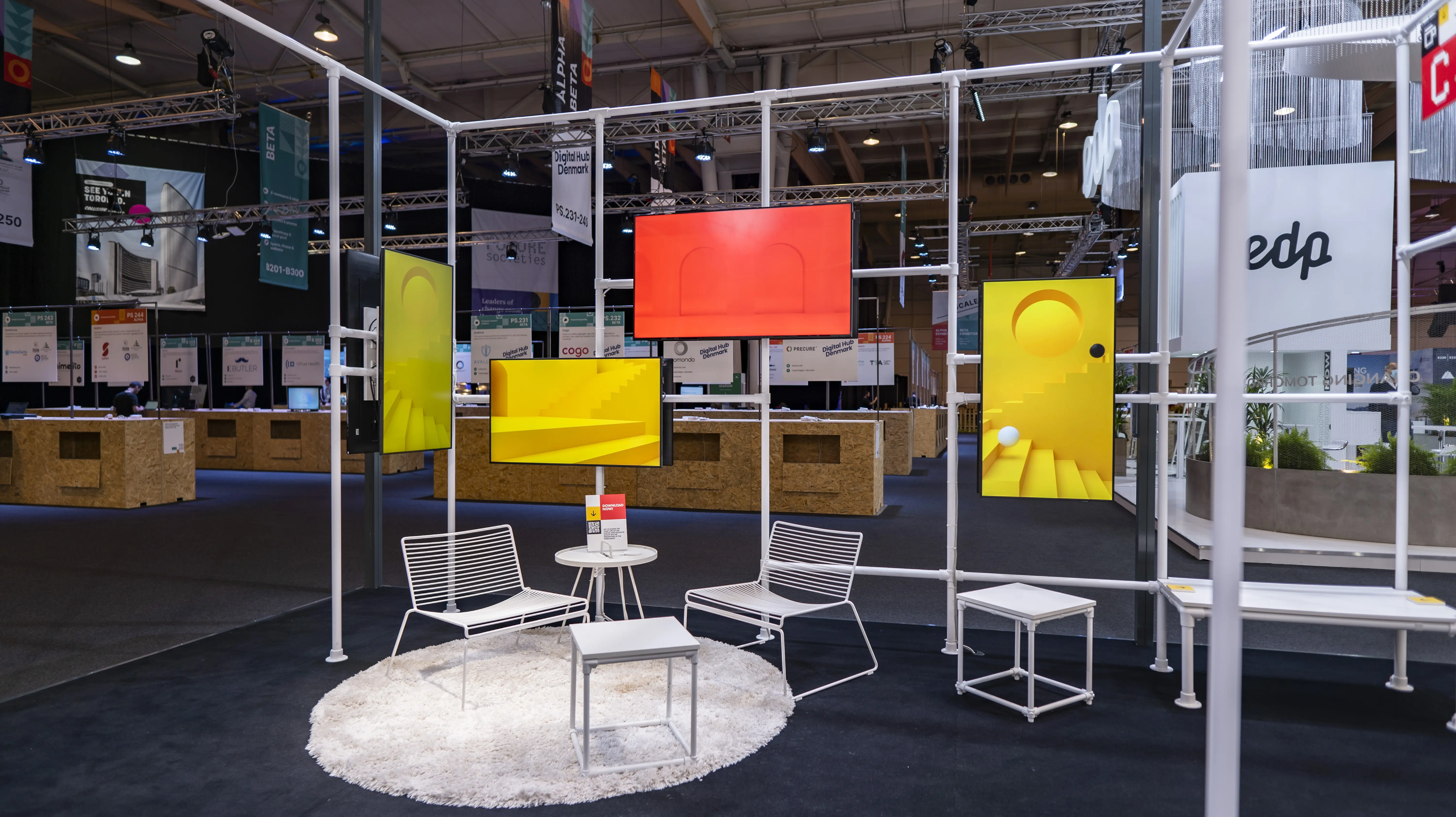 Lounge corner with white wire chairs on a round rug under colorful screens at the German Pavilion at Web Summit.