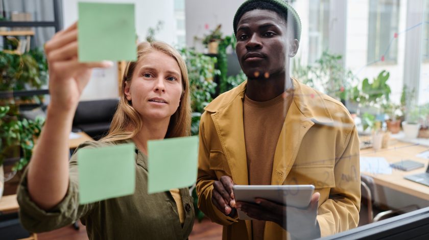 Two colleagues, one holding a tablet, collaborate on a project by placing green sticky notes on a glass interior office wall, surrounded by modern furniture and lush greenery.