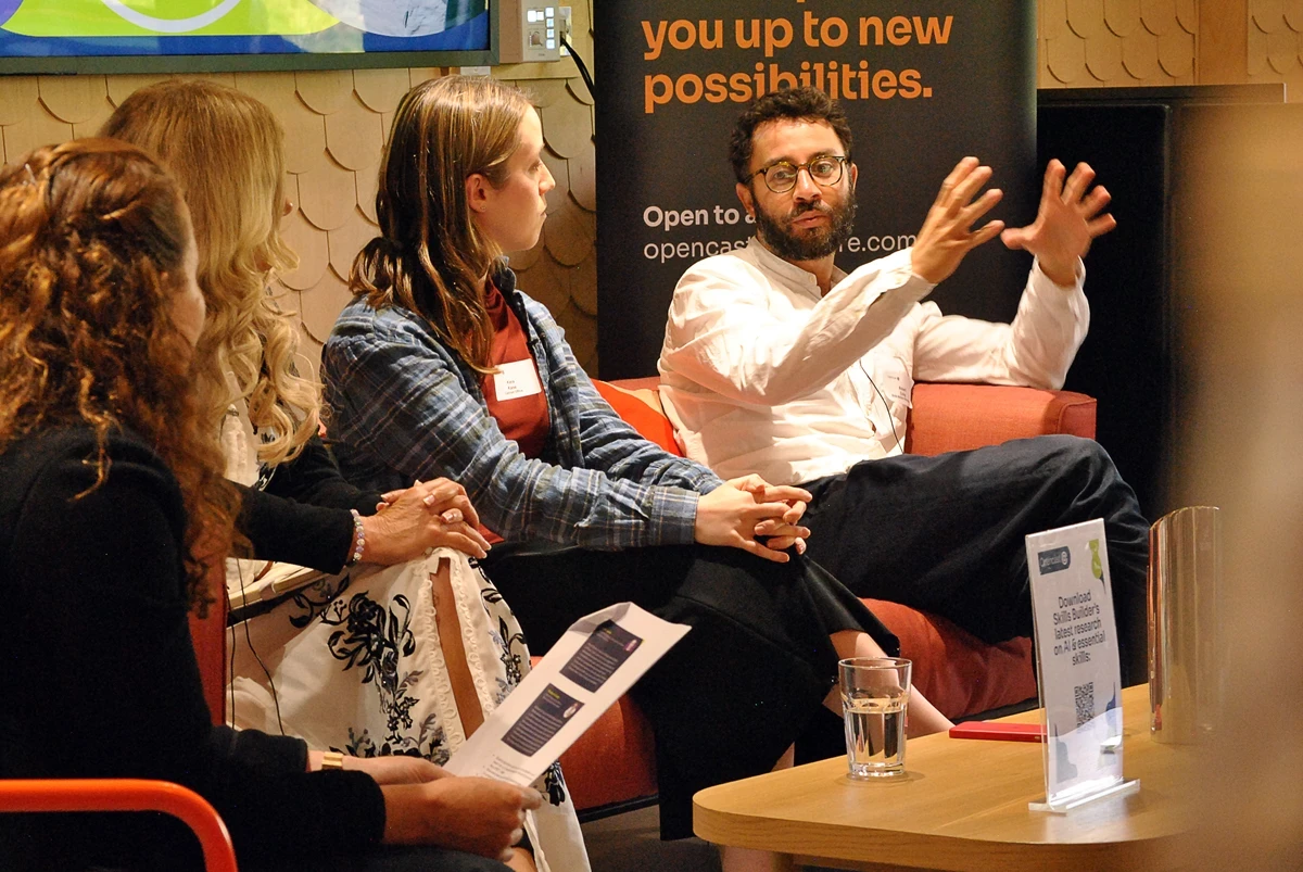 Panel discussion with four people seated on chairs and a sofa, one person speaking with hands raised. A banner in the background reads “you up to new possibilities.” A table in front holds a glass of water and a sign.