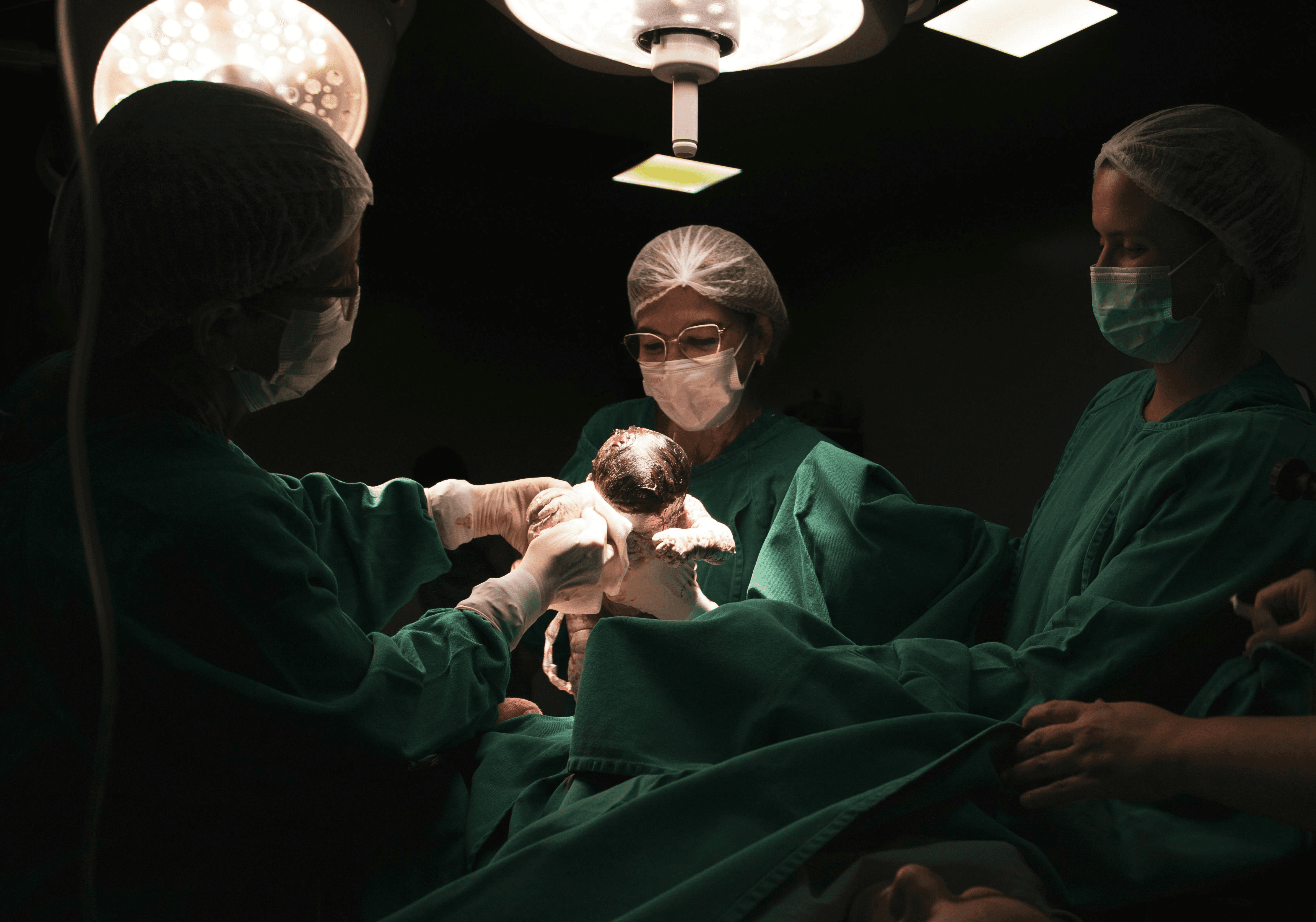 A dark operating theatre with a spot light on the focal point. A pair of doctors in turquoise scrubs, white masks and hair nets lifts a newborn up as part of the caesarean section procedure.