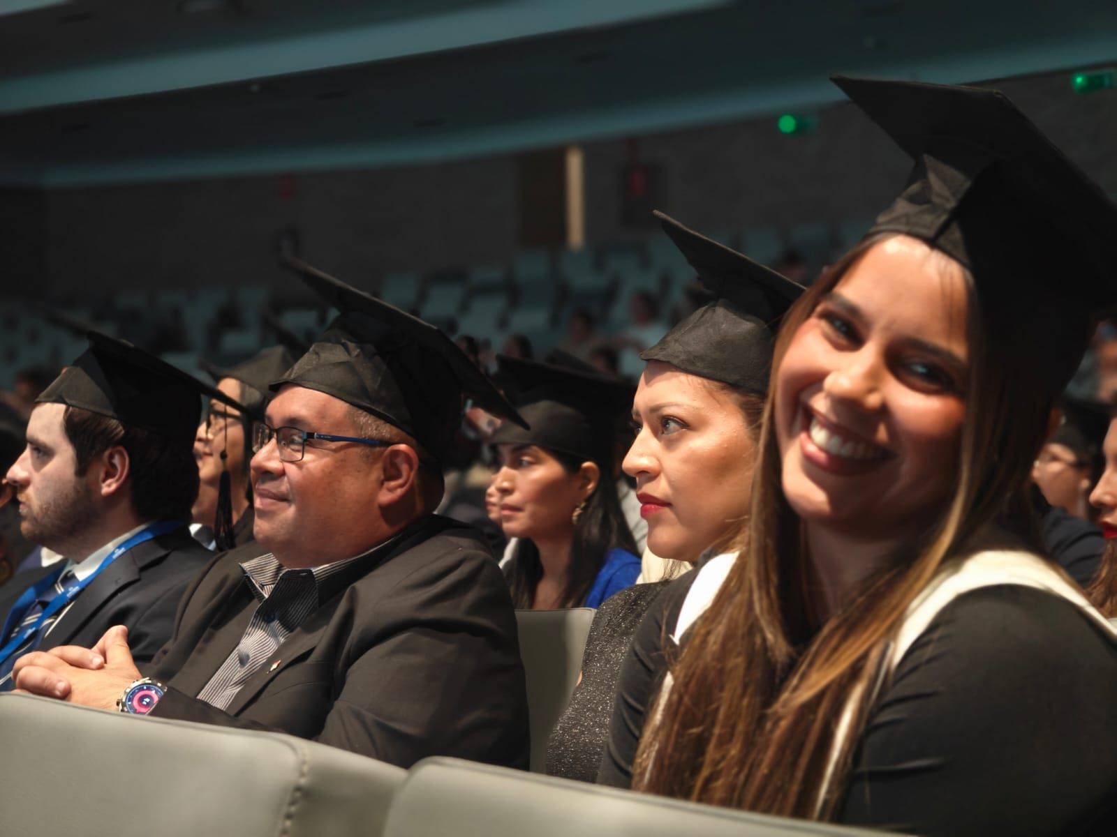 Young man in a green hoodie sitting at a table with a yellow phone in hand and a tablet in front of him, smiling at the camera.