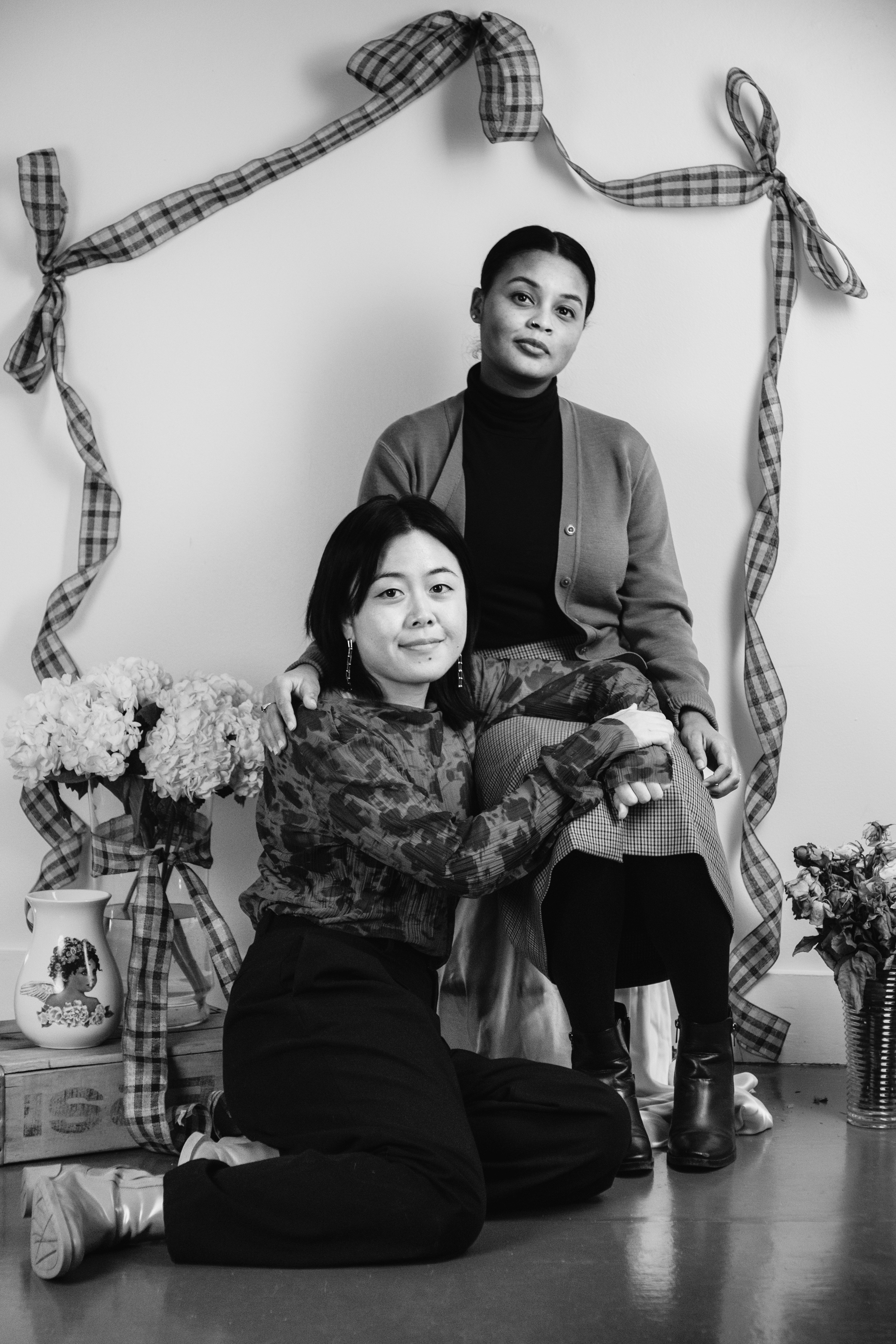 Attia and Kaori, two people pose in a monochrome photo, framed by plaid ribbons; one sits on a chair, while the other kneels beside them with floral arrangements and a decorative vase in the background, creating a stylish, vintage-inspired scene.