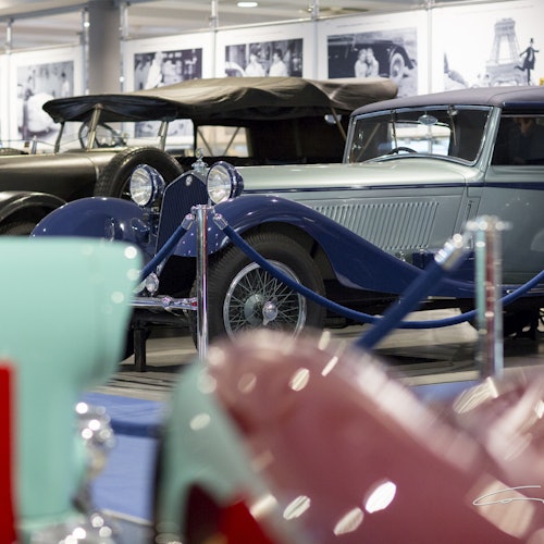 اتومبیلهای قدیمی در یک نمایشگاه به نمایش گذاشته شدهاند و طنابهای آبی آنها را جدا کردهاند. عکسهای تاریخی در پسزمینه قرار دارند.