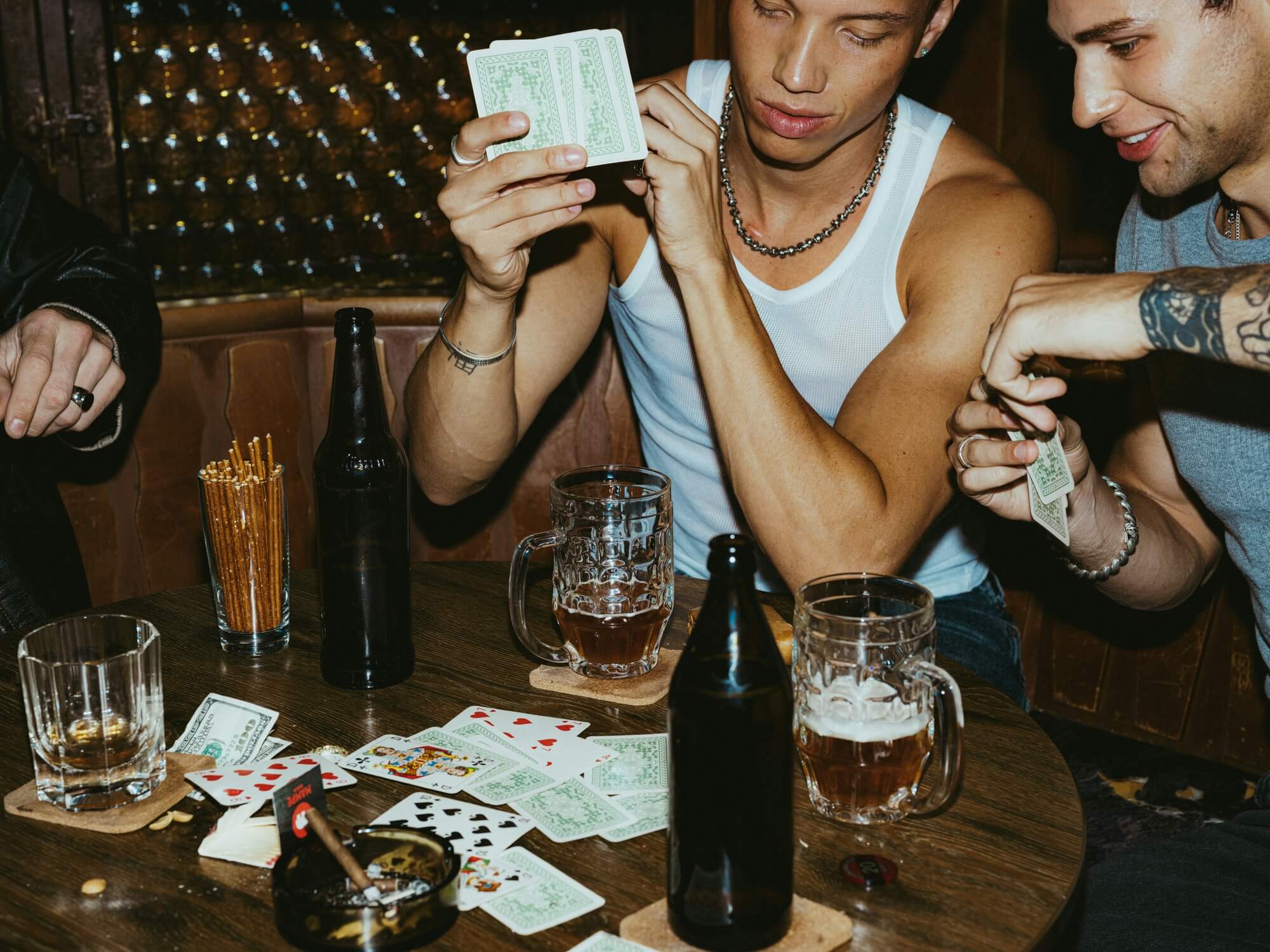 A group of people playing poker at a dimly lit bar table, with scattered cards and beers. The scene feels lively and social, focused on a man holding cards.