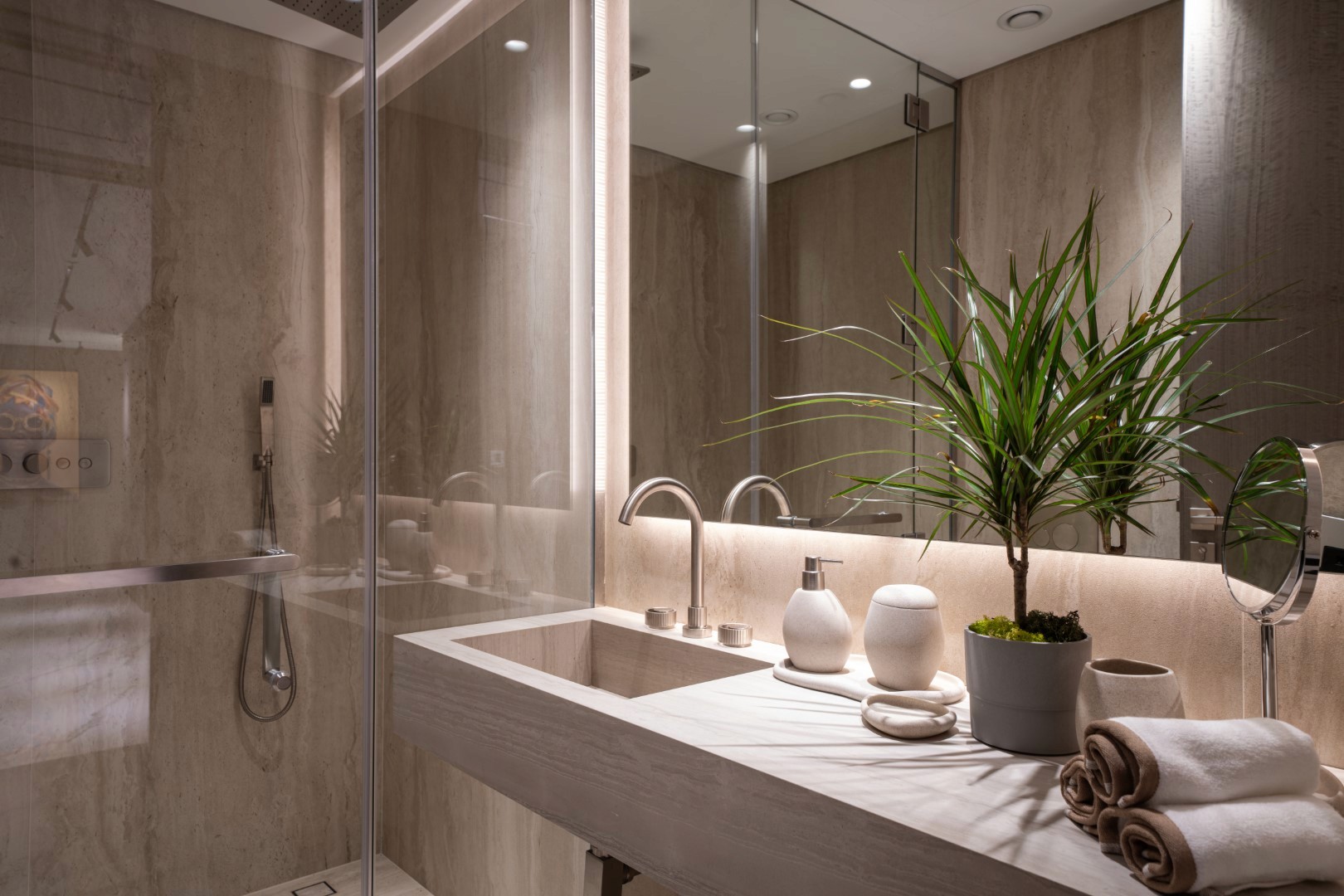 A modern bathroom featuring a sleek, gray stone countertop with a built-in sink, complemented by elegant faucet fixtures and enhanced by warm lighting; nearby, a vibrant green potted plant, a neatly rolled towel, and stylish toiletry containers sit beside a round mirror, while a walk-in glass shower enclosure adds a touch of luxury and sophistication.