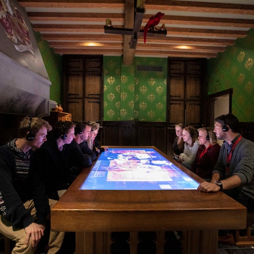 Emberek ülnek egy nagy interaktív asztal körül egy félhomályos szobában, fa gerendákkal és zöld mintás falakkal, fejhallgatót viselve.