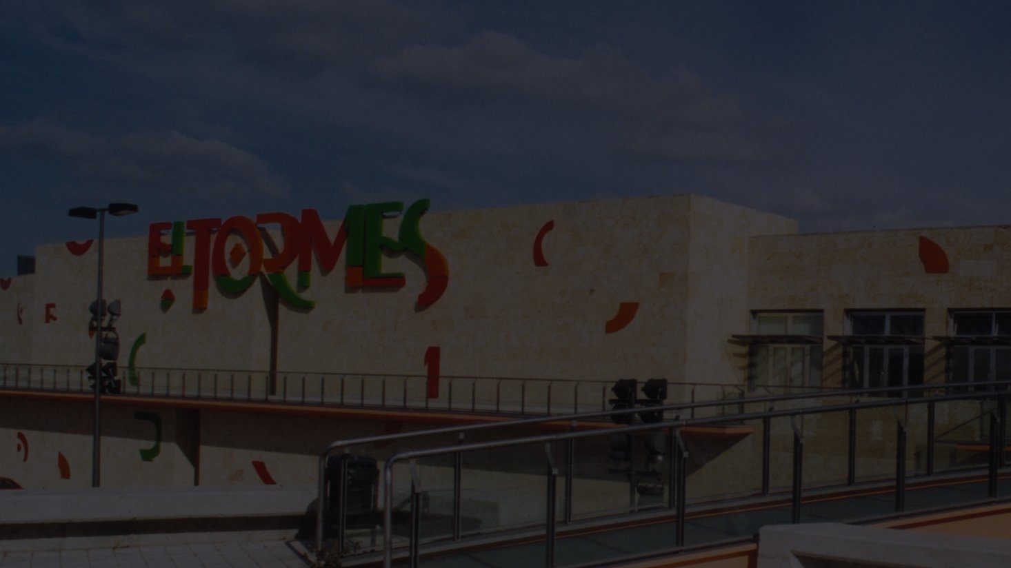 Modern shopping center exterior with colorful "El Tormes" signage atop a beige stone facade. Glass railing and scattered clouded sky add an inviting ambiance.