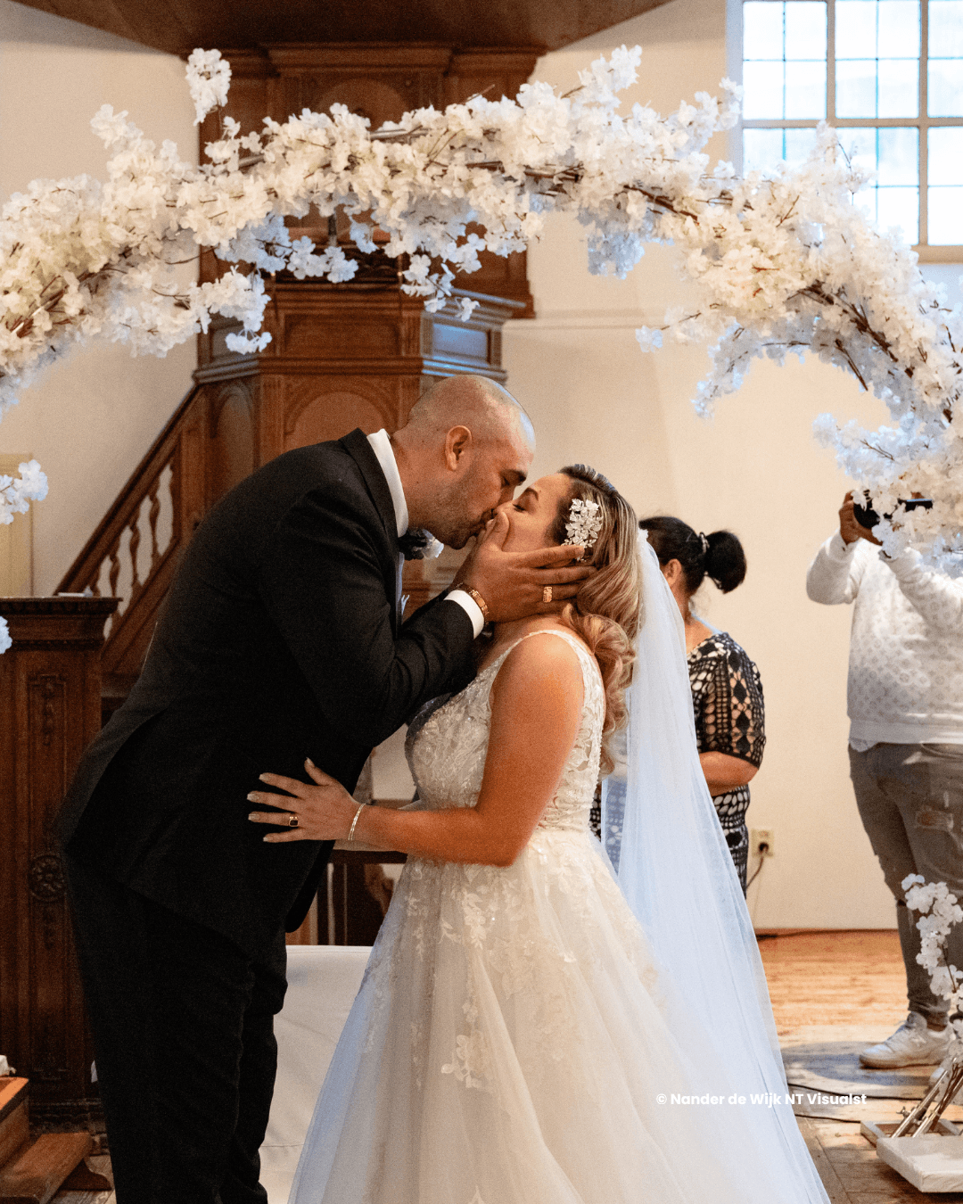 A couple in formal attire shares a kiss under an elegant floral arch during their wedding ceremony.