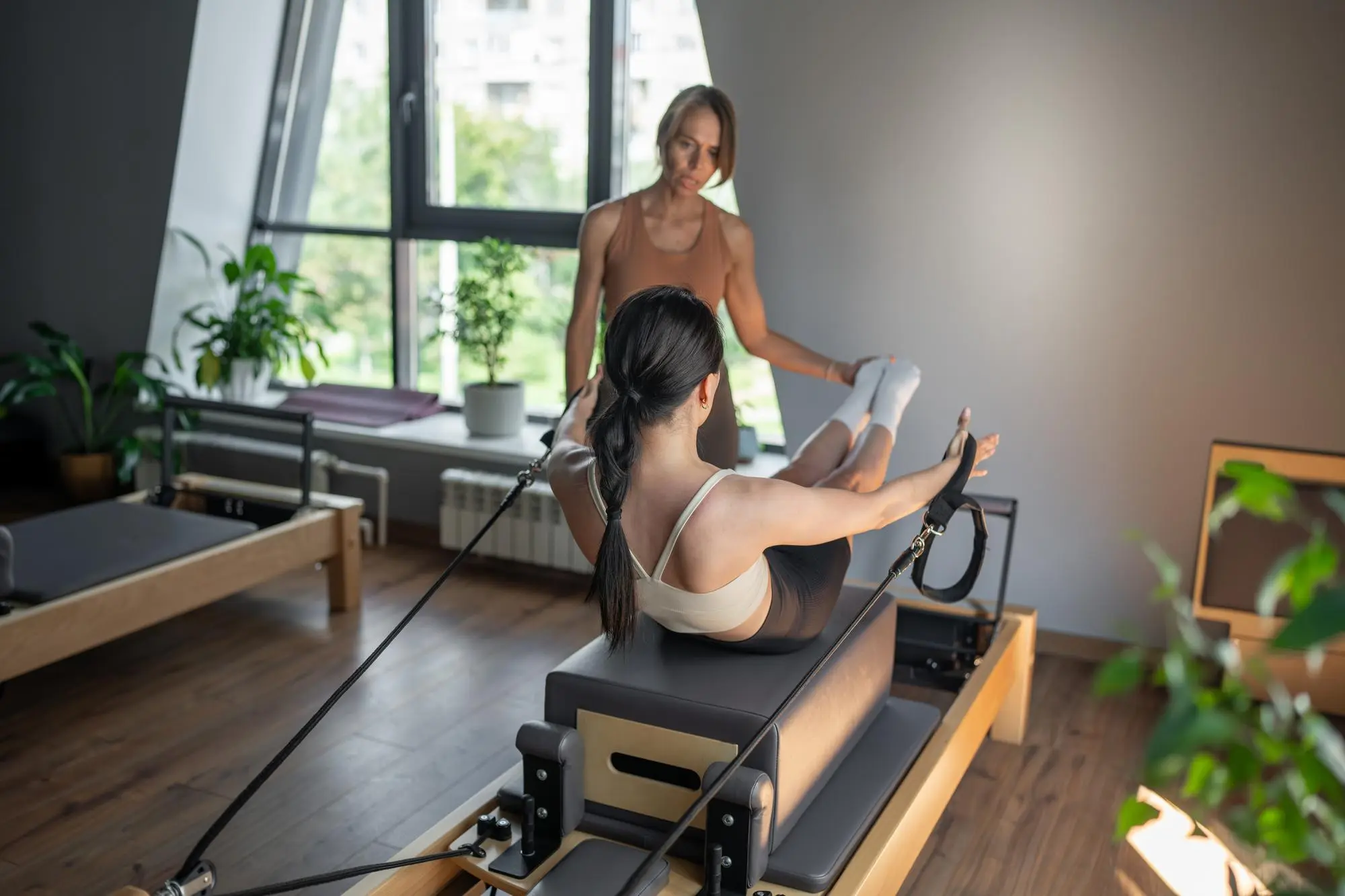 Two people are in a bright studio; one is assisting the other on a reformer Pilates machine.
