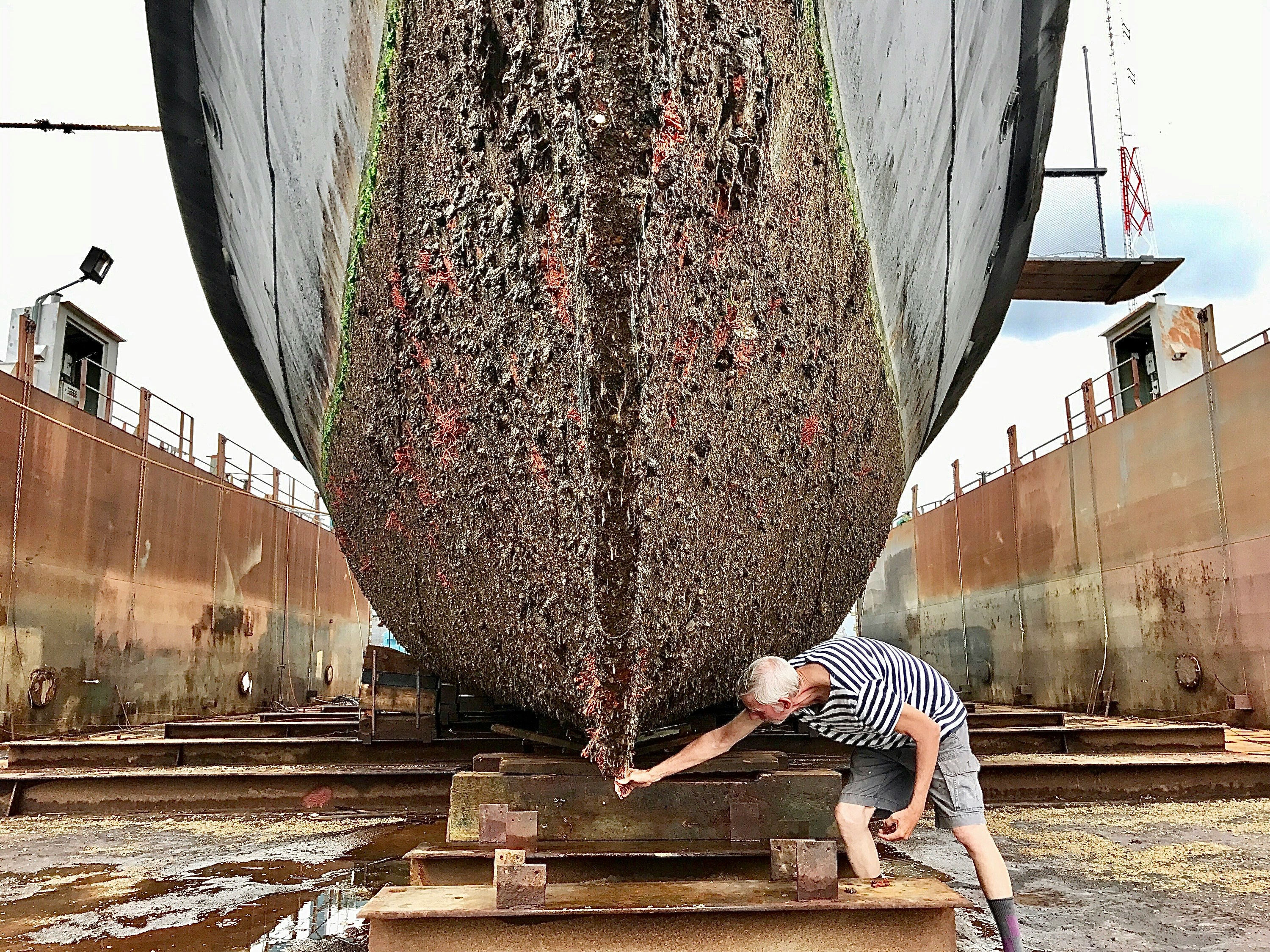 Heavily fouled boat hull covered in barnacles and marine growth in a dry dock with a man inspecting the keel