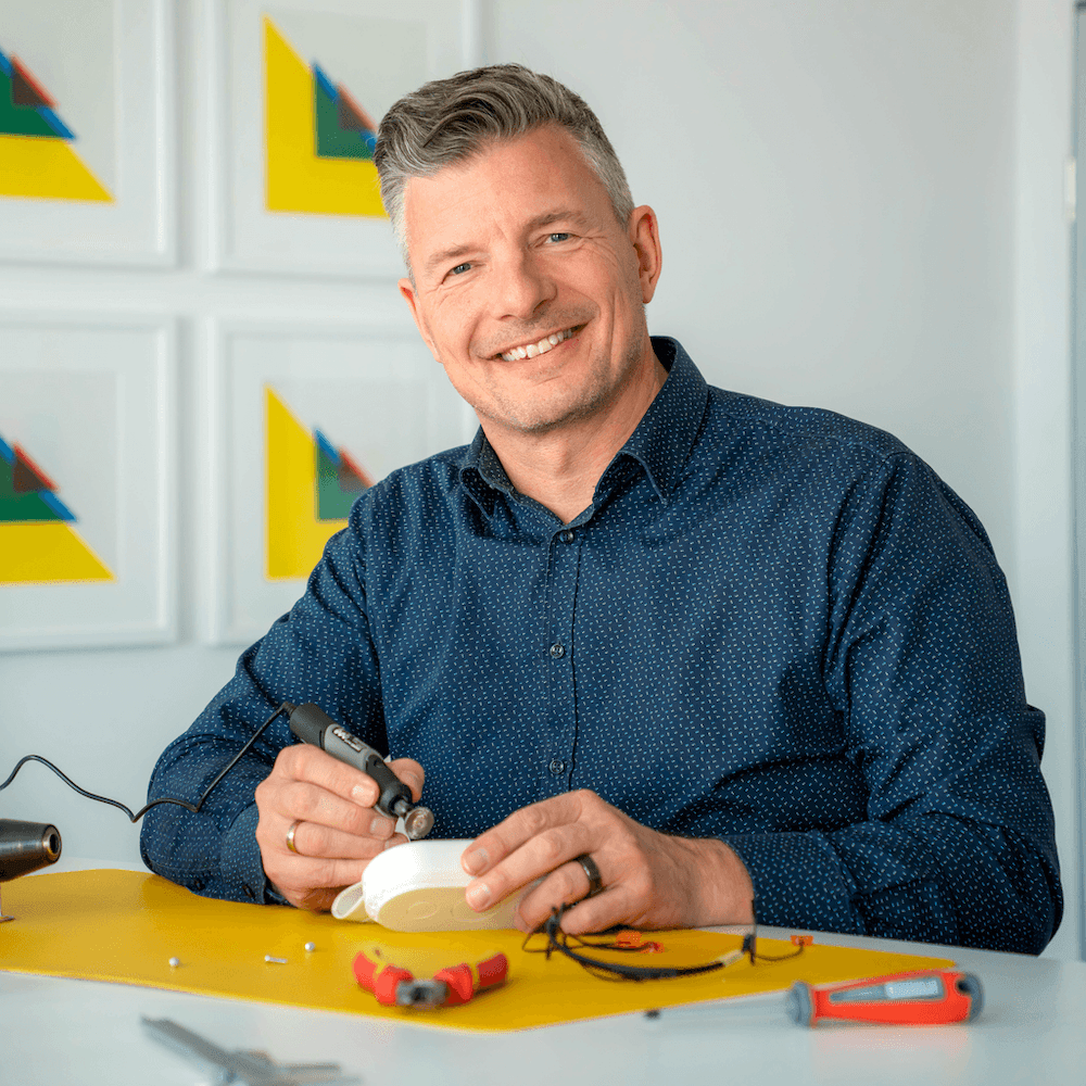 Mann mit blauem Hemd sitzt an einem Tisch und hält ein elektrisches Werkzeug über einem weißen Objekt