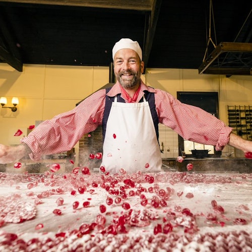一位戴着白色围裙和帽子的胡须男士站在桌子后面,微笑着在撒了粉的表面上处理红色糖果。