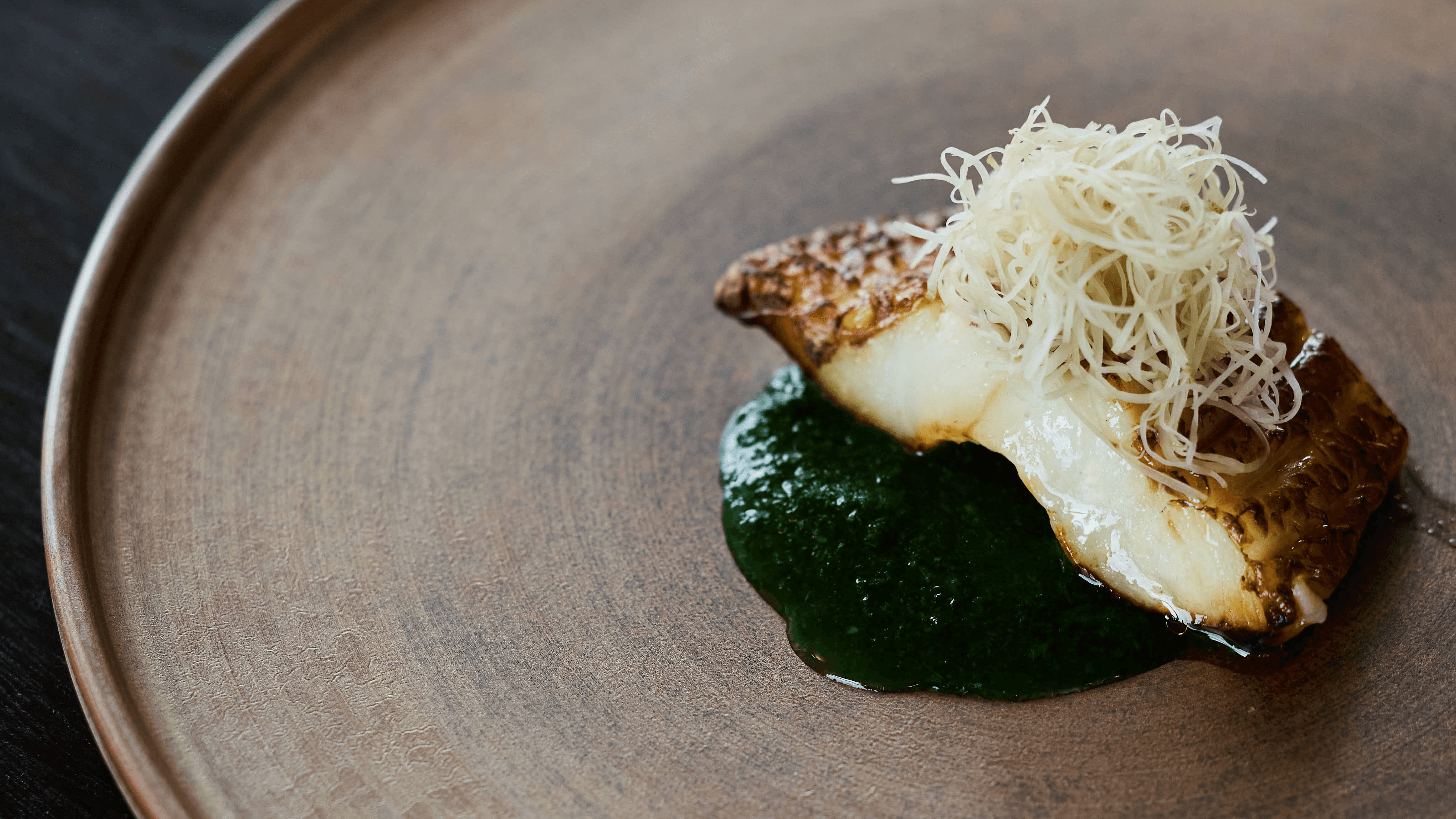 A masterfully plated Japanese Kappo dish featuring a piece of fire-grilled fish resting on a vibrant green herb puree, topped with delicate white garnishes and served on a textured, earth-toned ceramic plate.